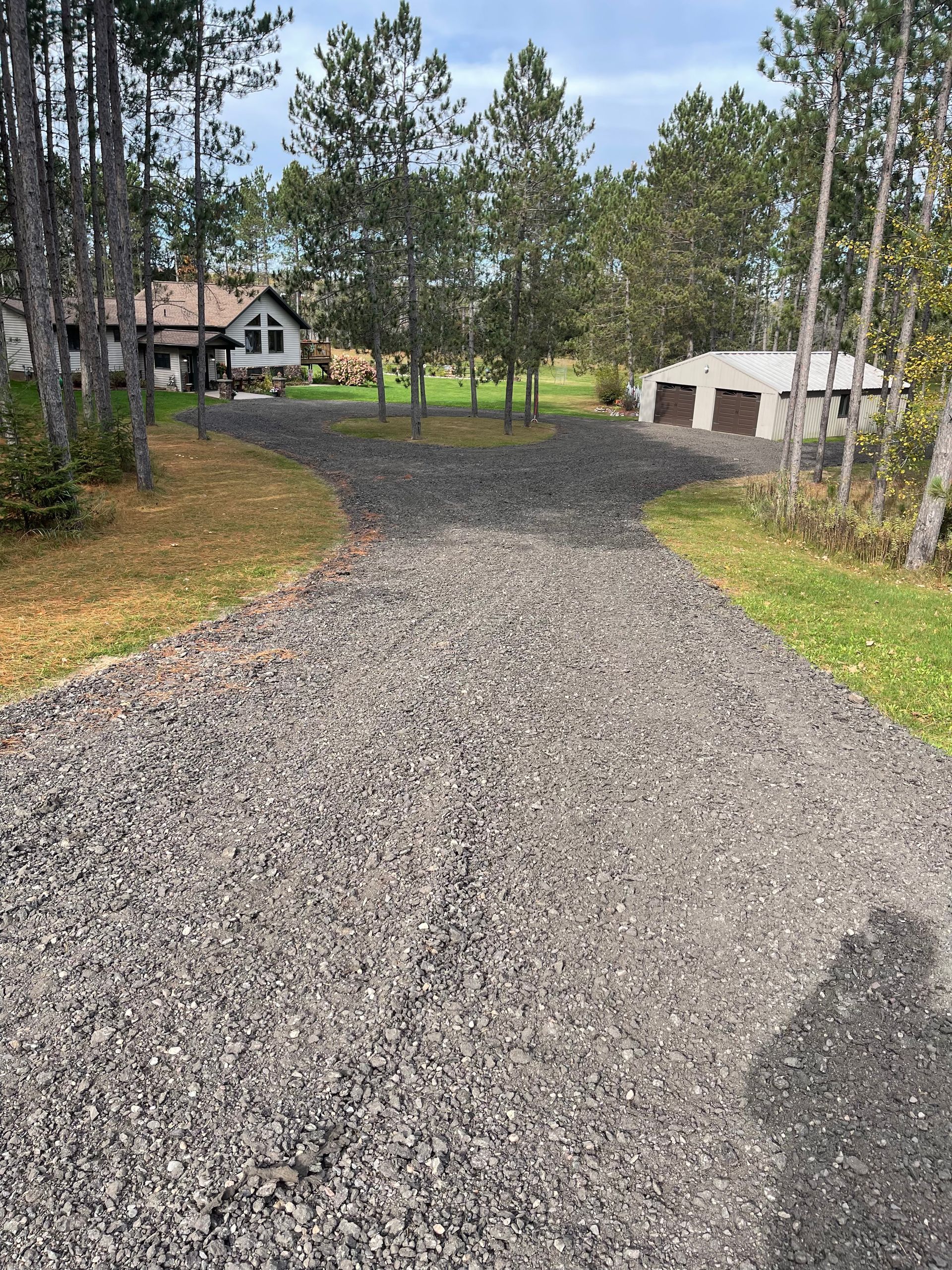 Gravel driveway forks, leading to a house and garage, surrounded by trees and grass, on an overcast day.