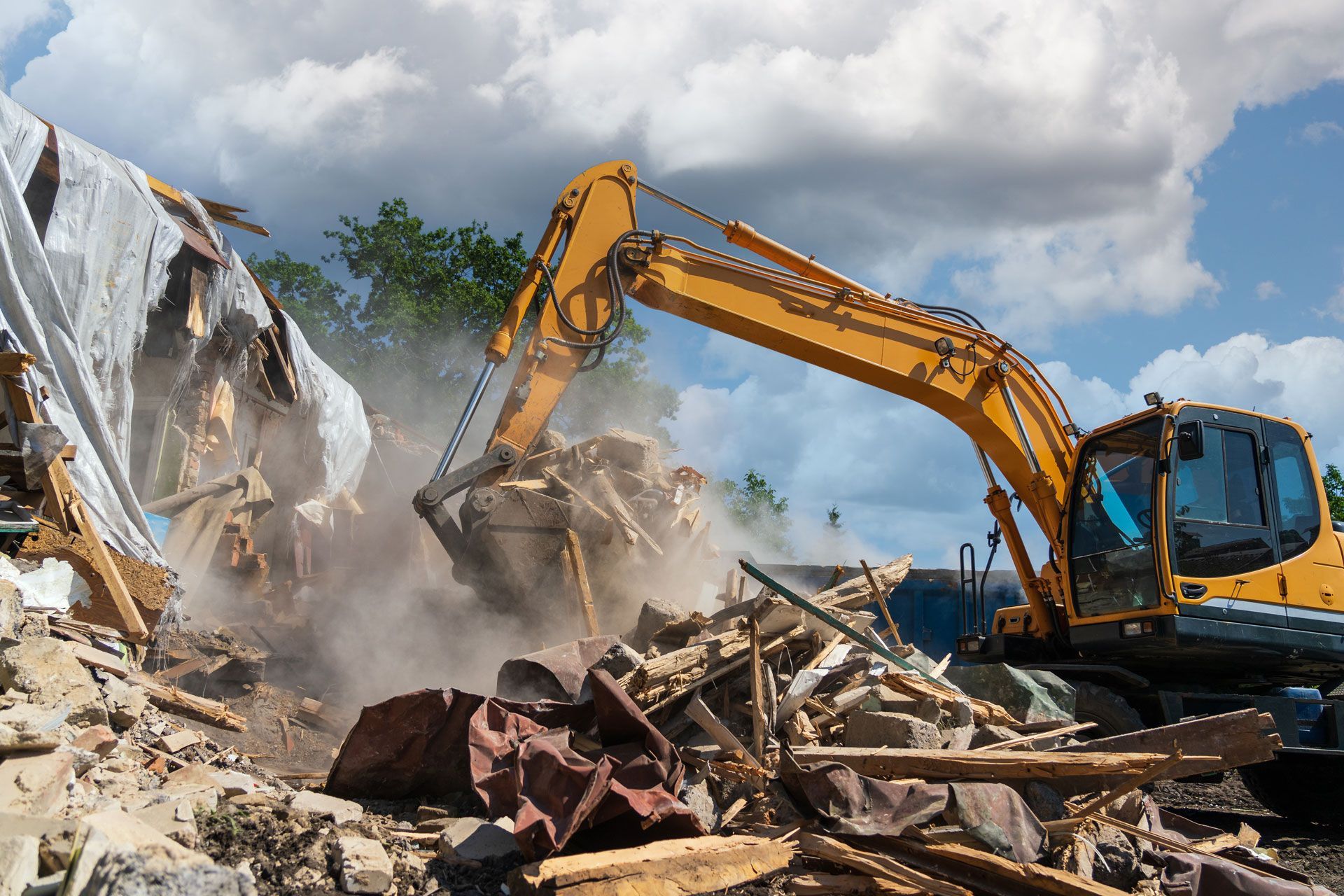 Yellow excavator demolishing a building; dust clouds in sunny, blue sky.
