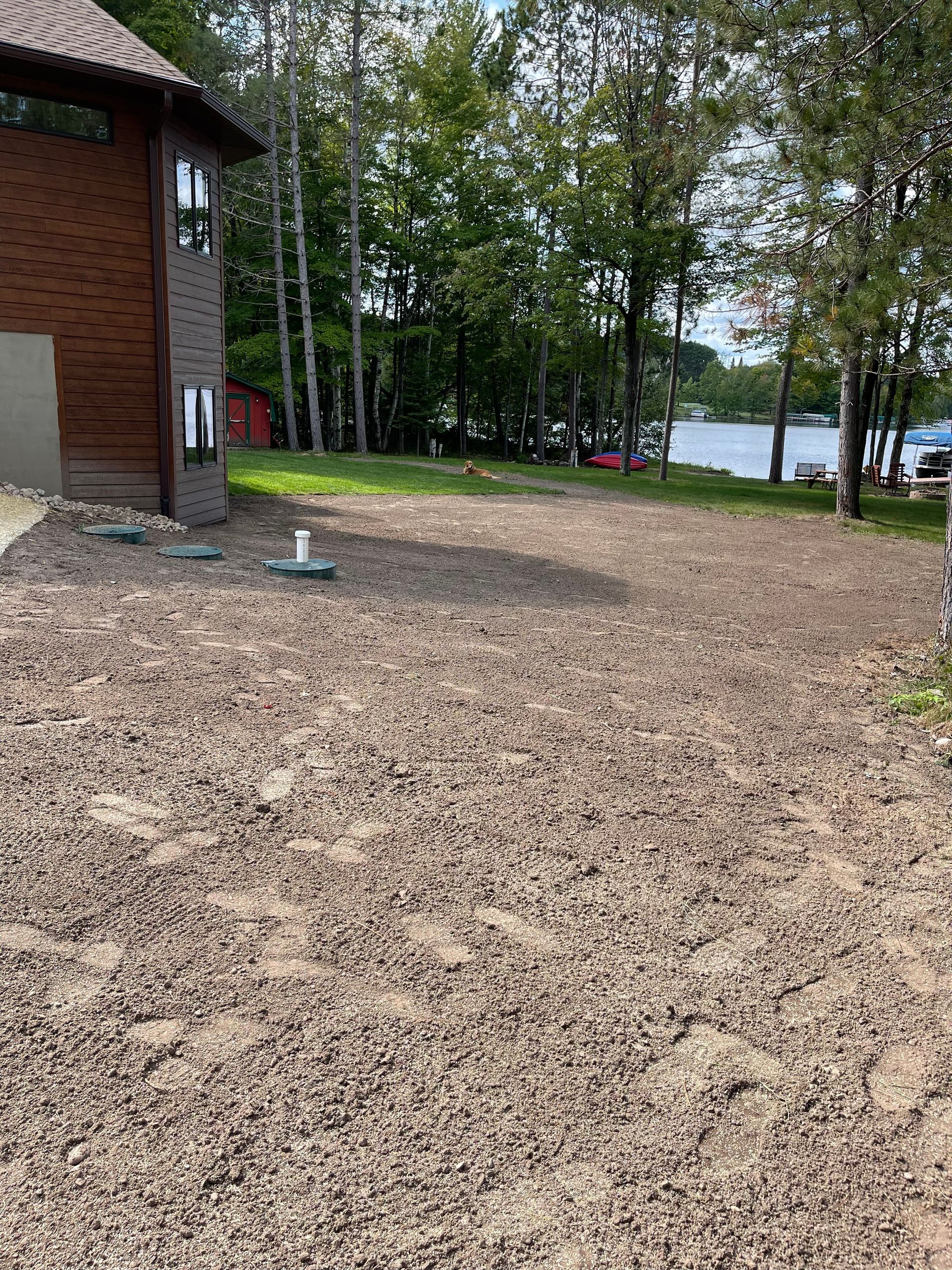 A cleared area of dirt and gravel near a building and trees, with a lake visible in the background.