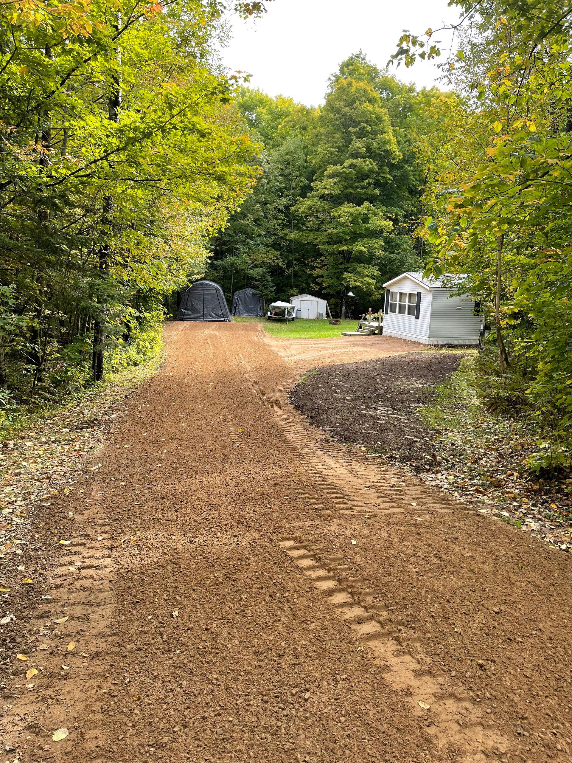 Dirt road leads to a wooded area with trees and a small trailer.