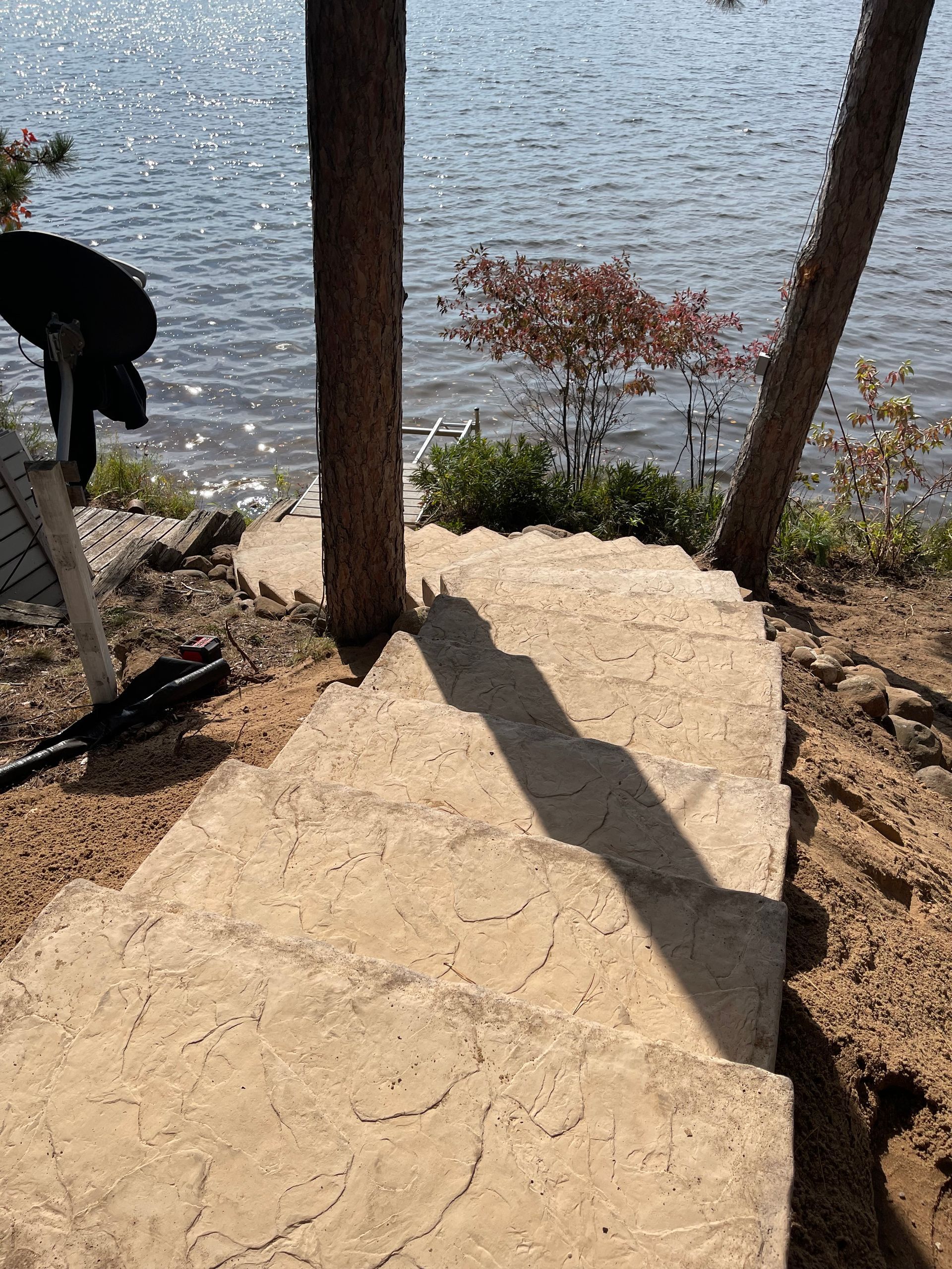 Concrete steps descend towards a lake, flanked by trees. Sunlight casts a long shadow on the steps.
