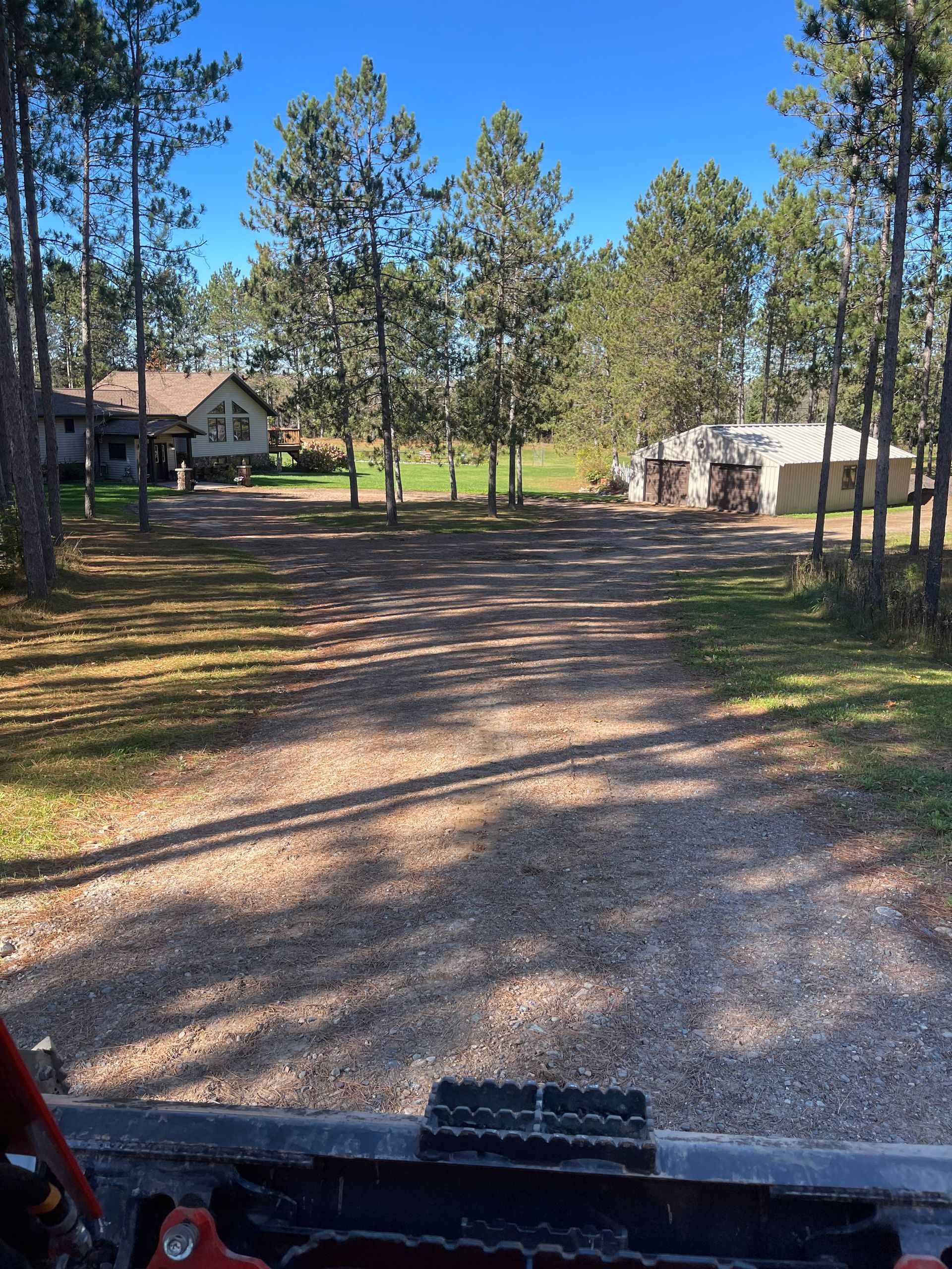 Gravel driveway leads toward house and garage on a sunny day. Tall trees line the sides.