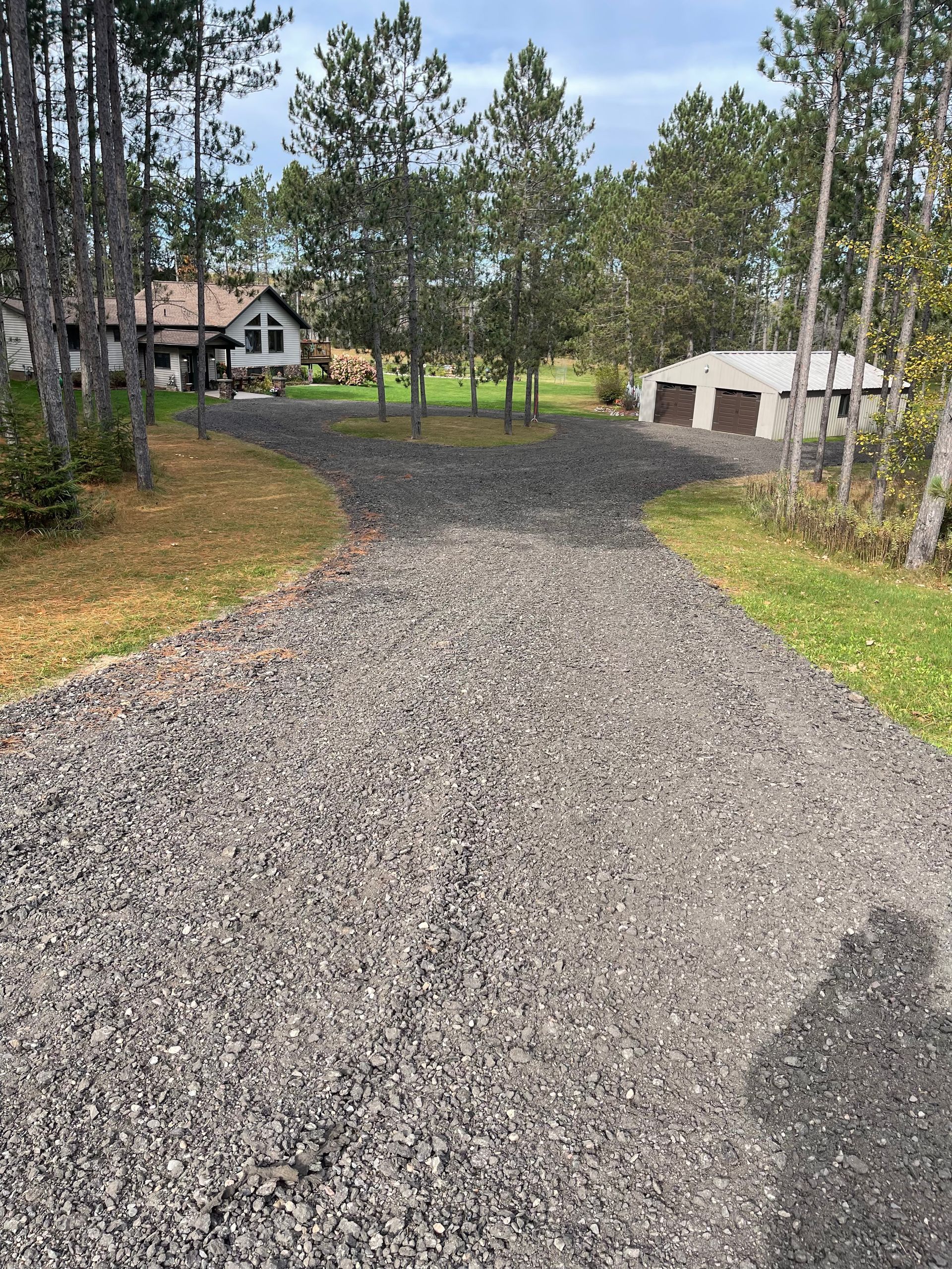 Gravel driveway splits into two paths, leading to a house and a garage, with trees in the background.