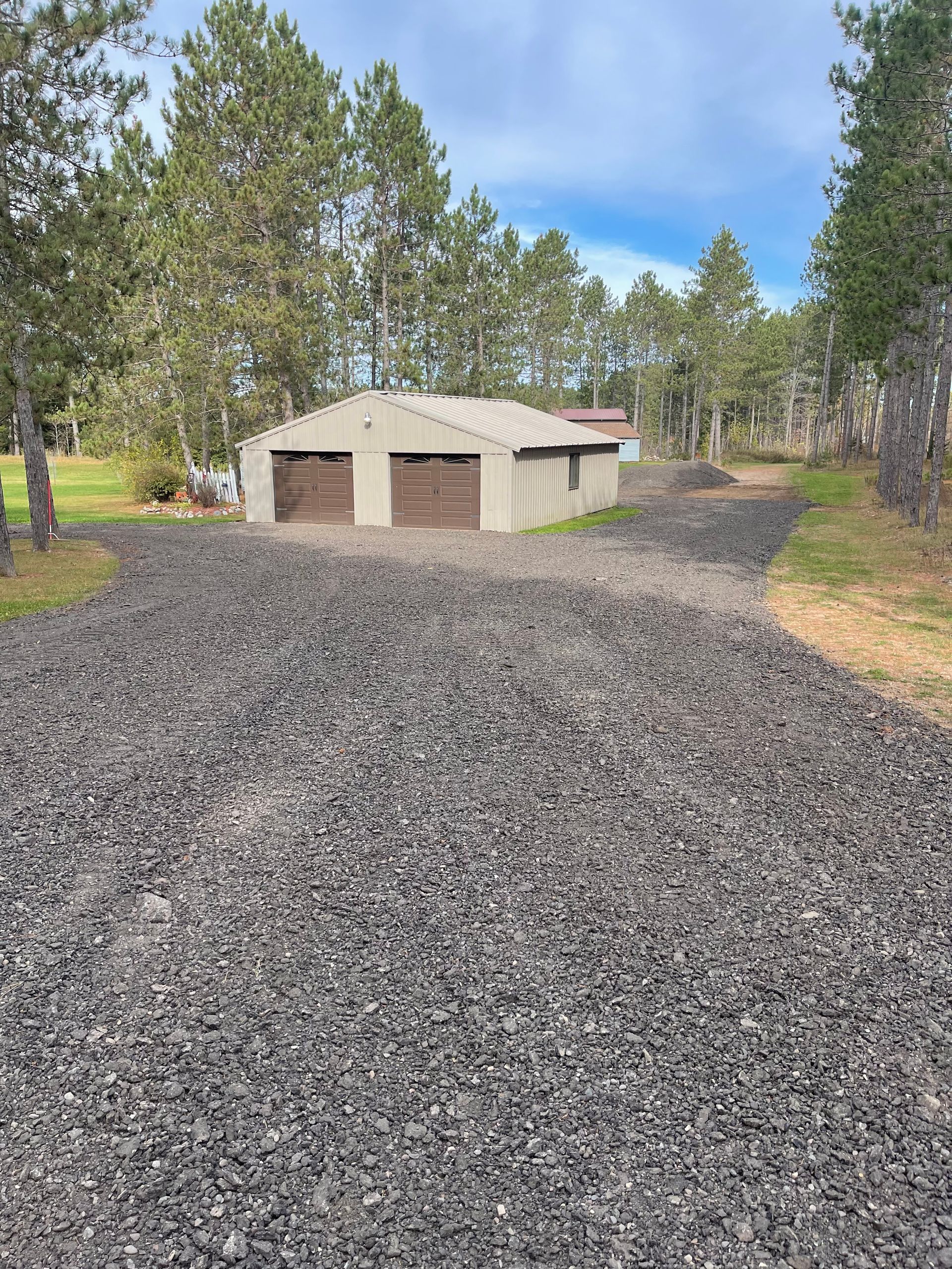 Gravel driveway leads to a two-car garage with brown doors, set in a wooded area under a cloudy sky.