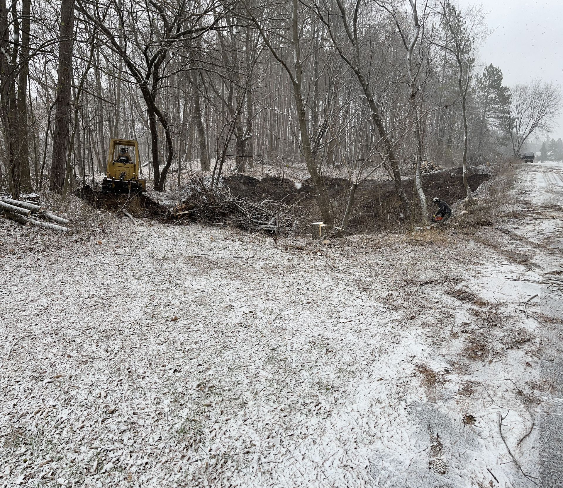Snowy scene with a yellow backhoe digging near a road lined by trees.