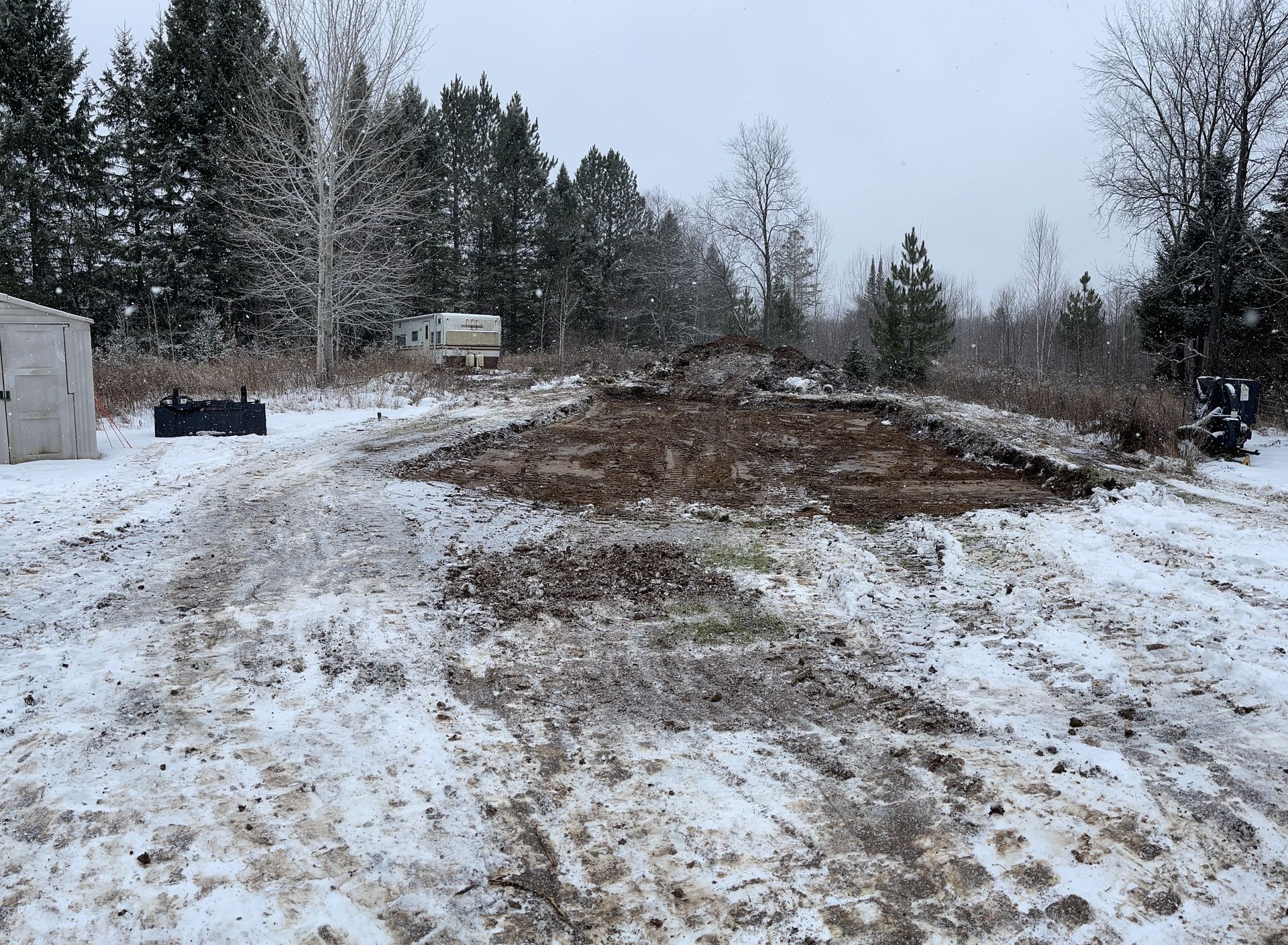 Snowy landscape with a dirt pile in the center and trees in the background. Construction equipment is also visible.