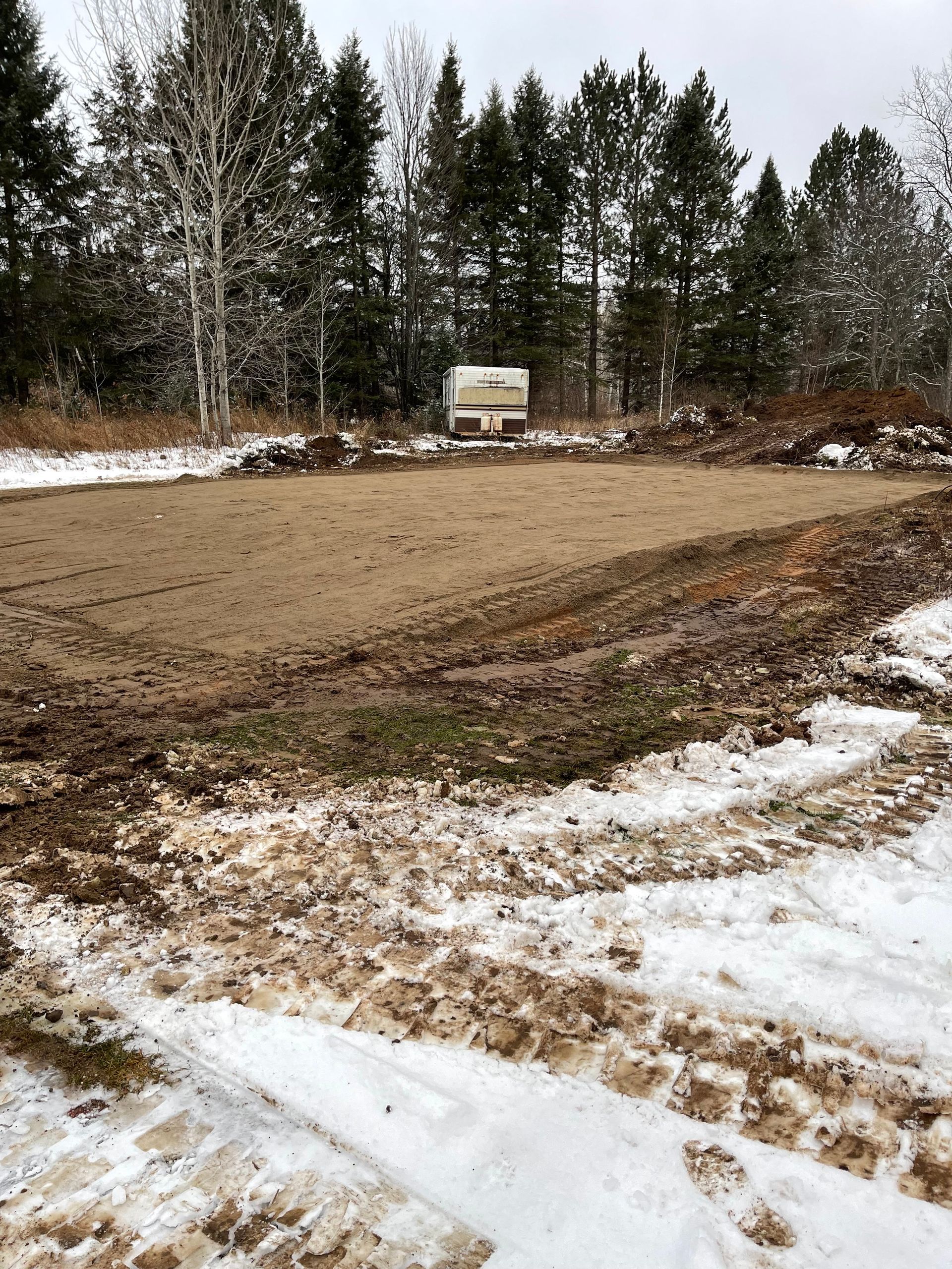 Snowy ground with muddy tire tracks leads to a leveled patch of dirt; a camper sits in the distance, backed by trees.