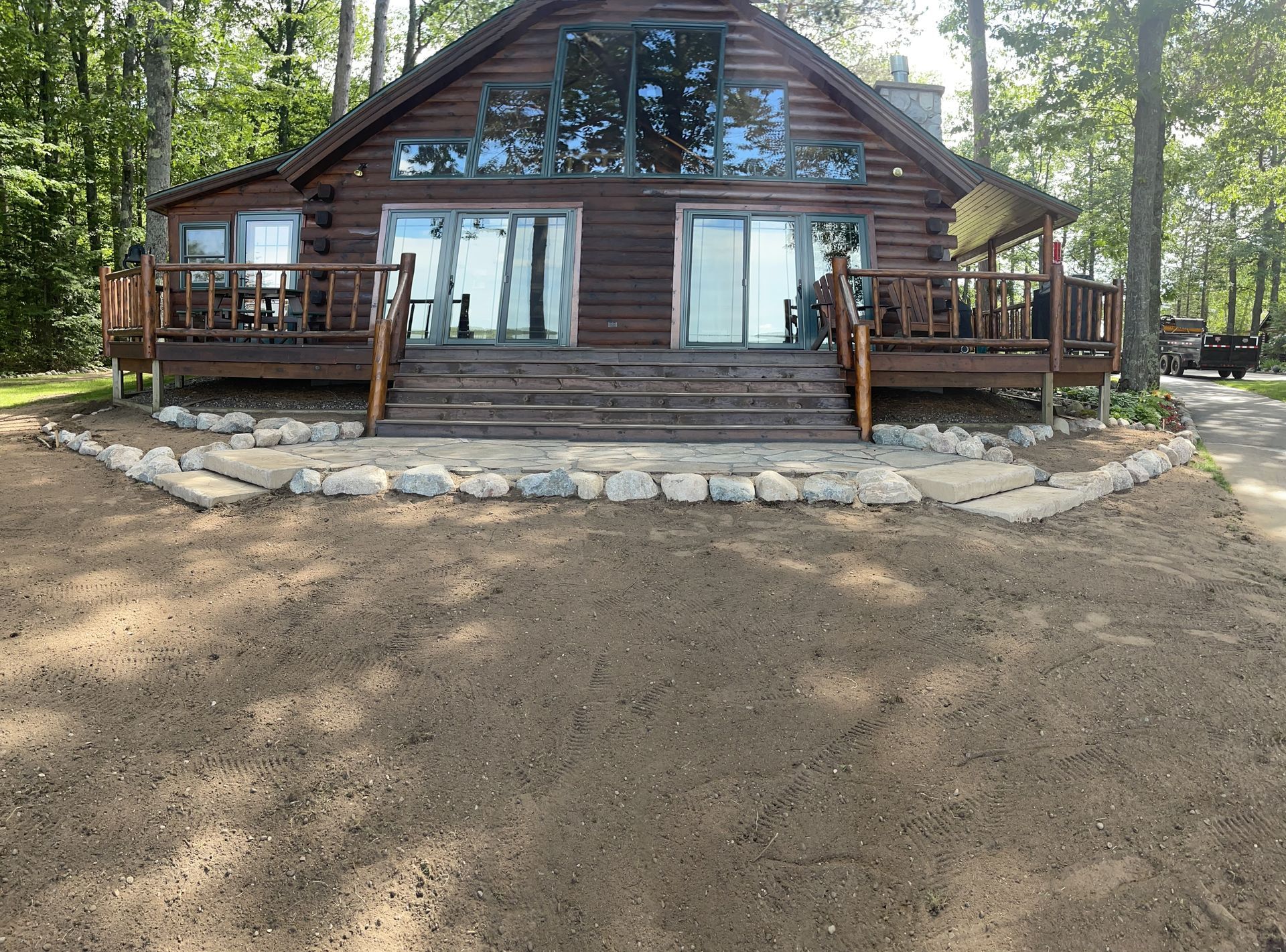 Log cabin with large windows and a deck, facing a gravel area, set in a wooded area.