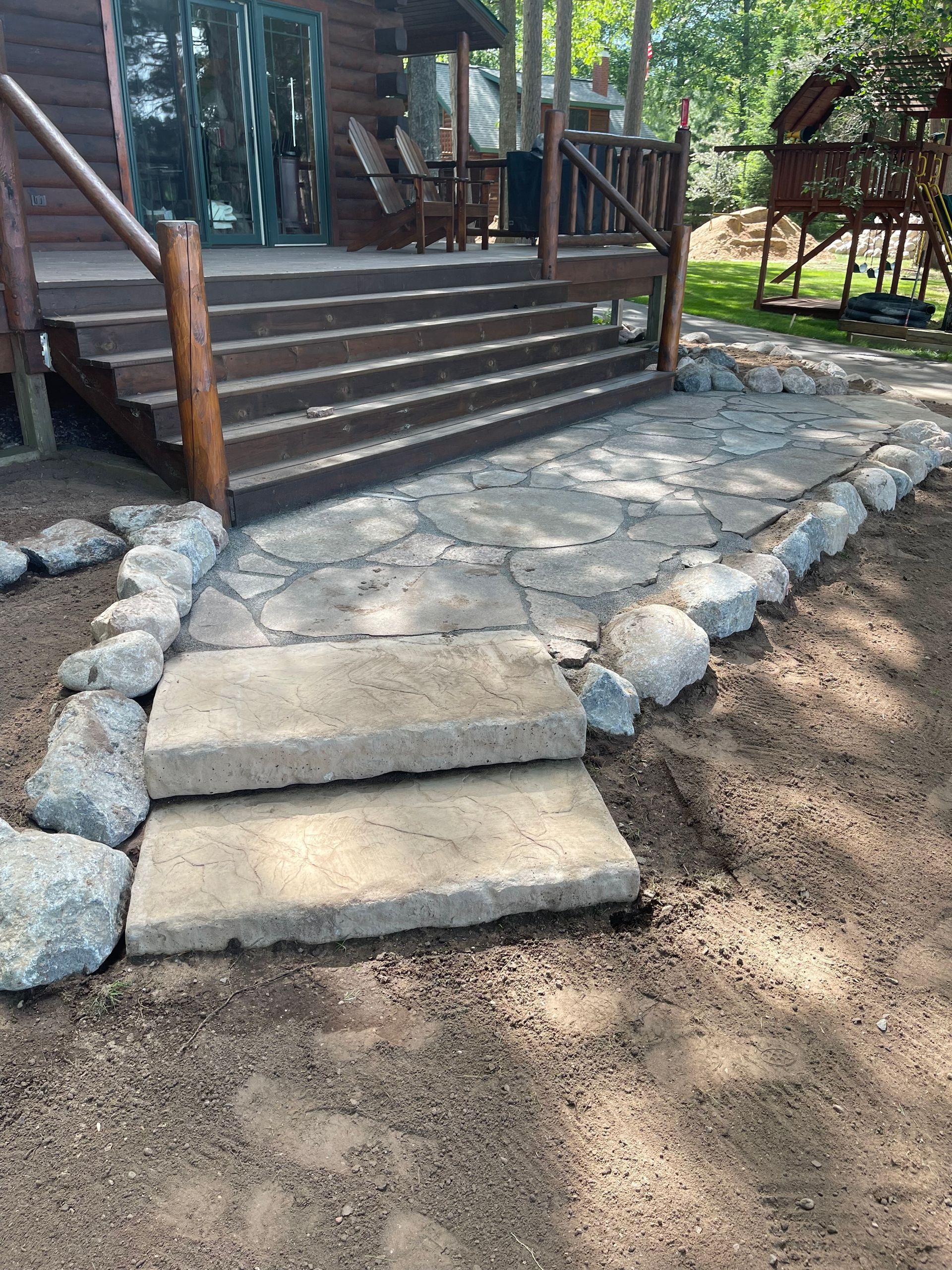 Stone patio with concrete steps leading to a wooden deck with railing. Large rocks border the patio.