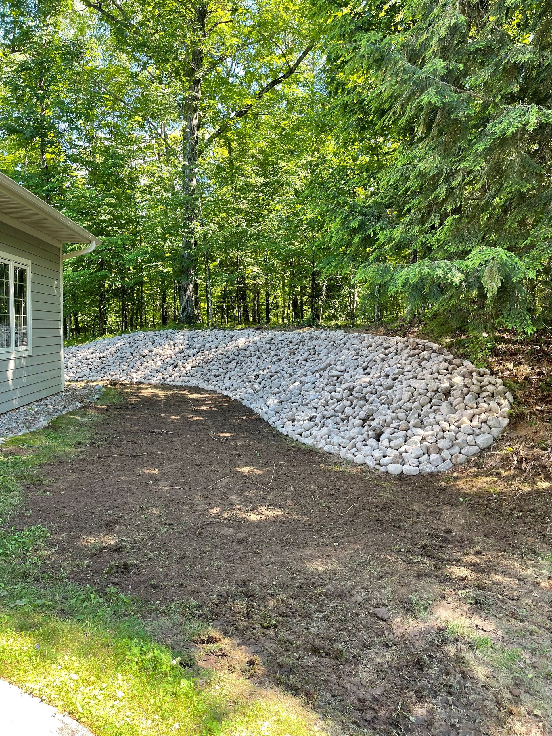 A curved rock garden bed next to a building, with exposed dirt in front, surrounded by grass and trees.