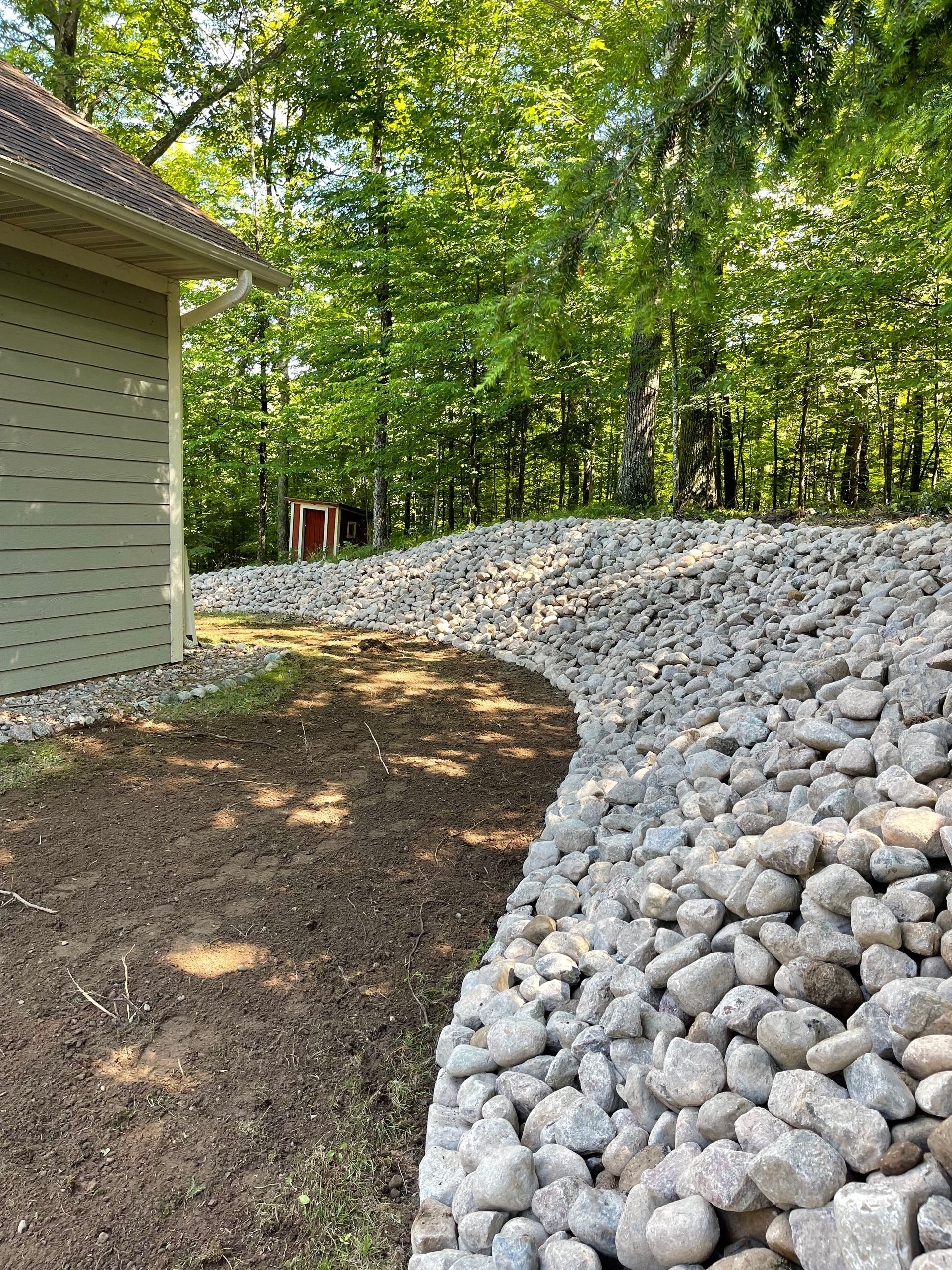 A curving rock retaining wall next to a shed and dirt patch, with a wooded background.