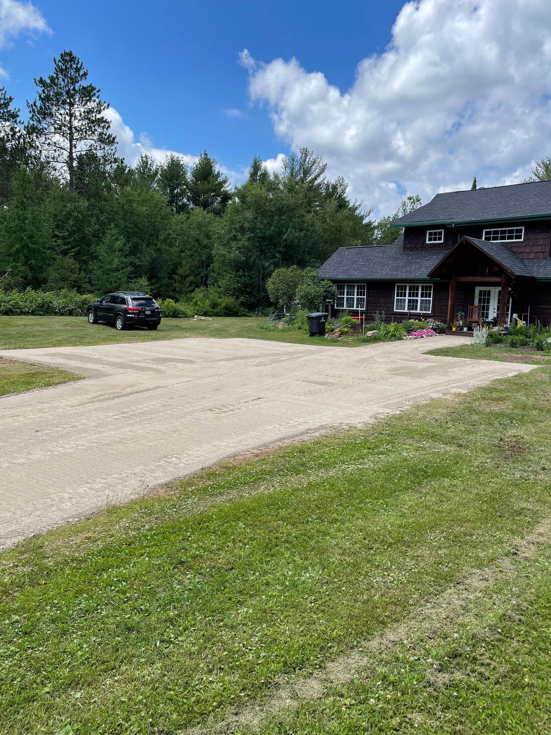 Gravel driveway leading to a brown house with a dark roof. A black car is parked. Green lawn and trees.
