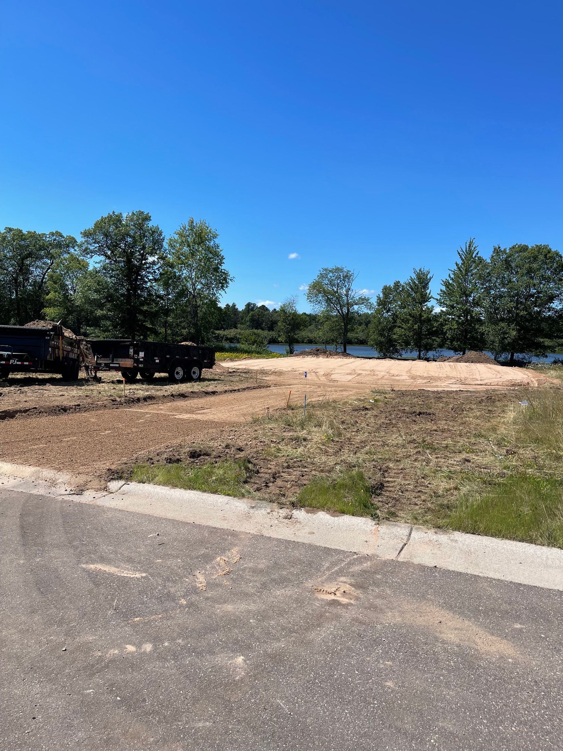 Cleared lot with bare dirt, trees, and blue sky; a body of water in the background.