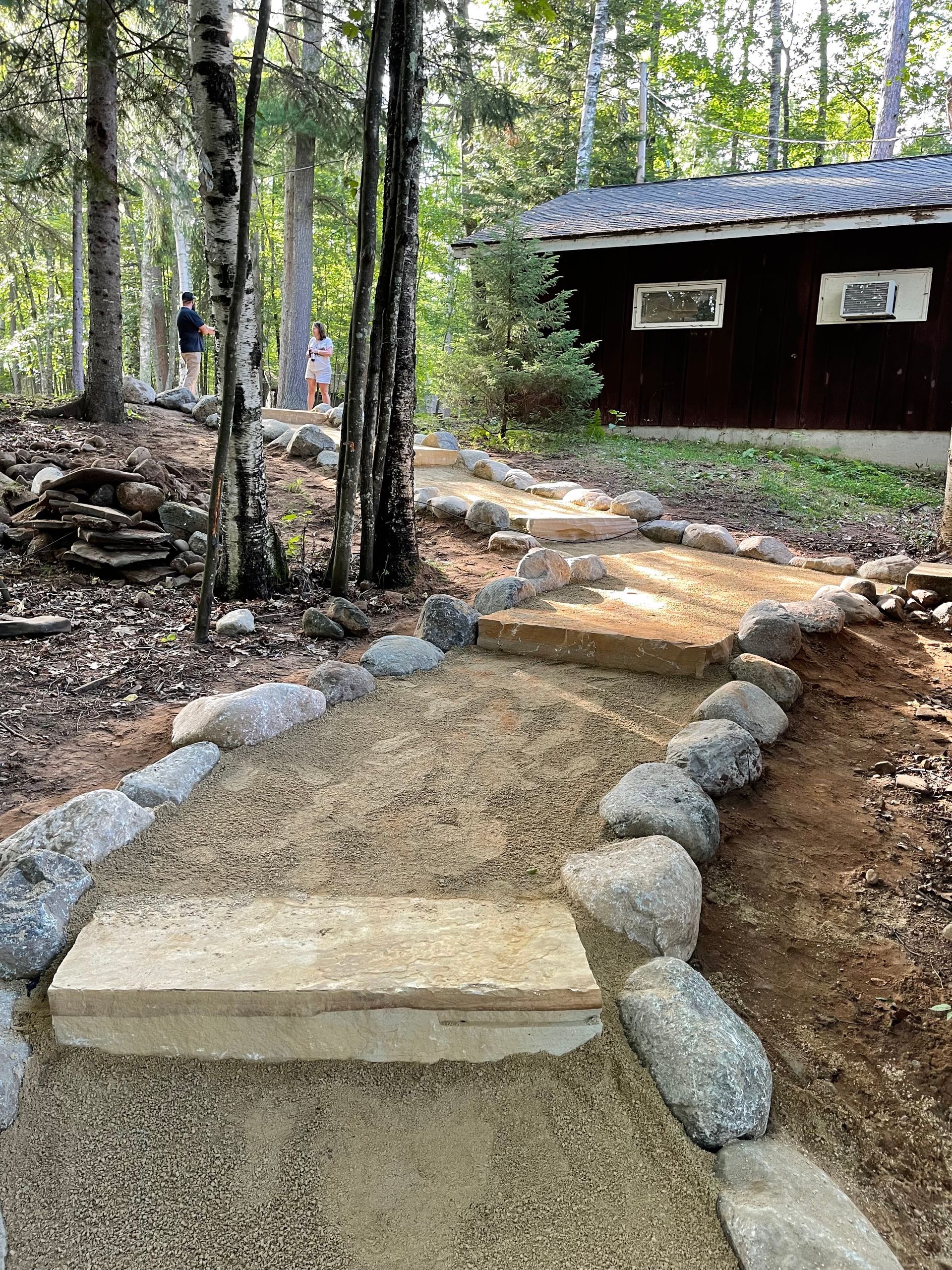 Stone steps lead up a sandy path bordered by rocks, near a dark building and trees.