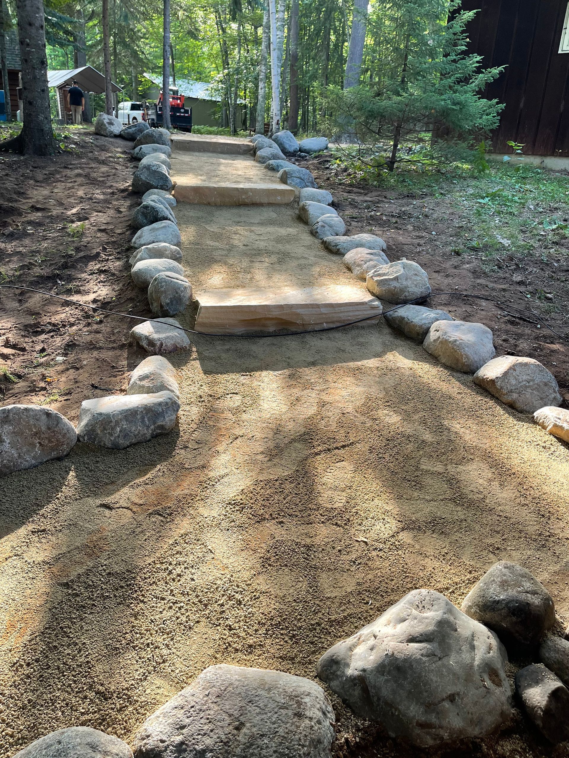 Stone pathway with wooden steps through a wooded area. The path is lined with large rocks.