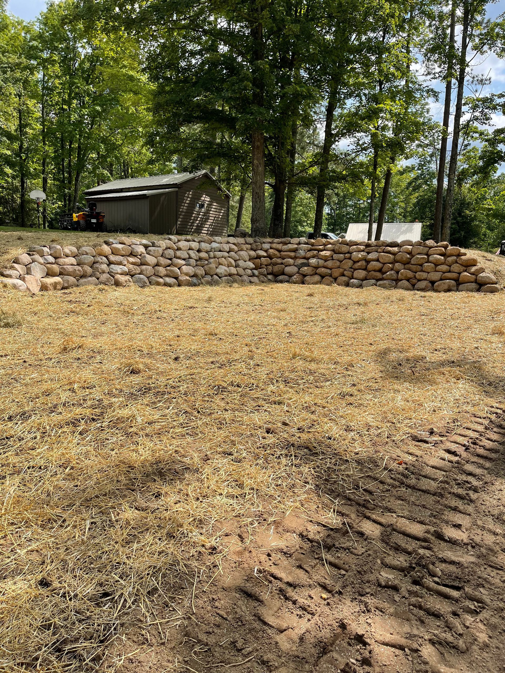 Brown hillside with stone retaining wall, small building, and trees in background.