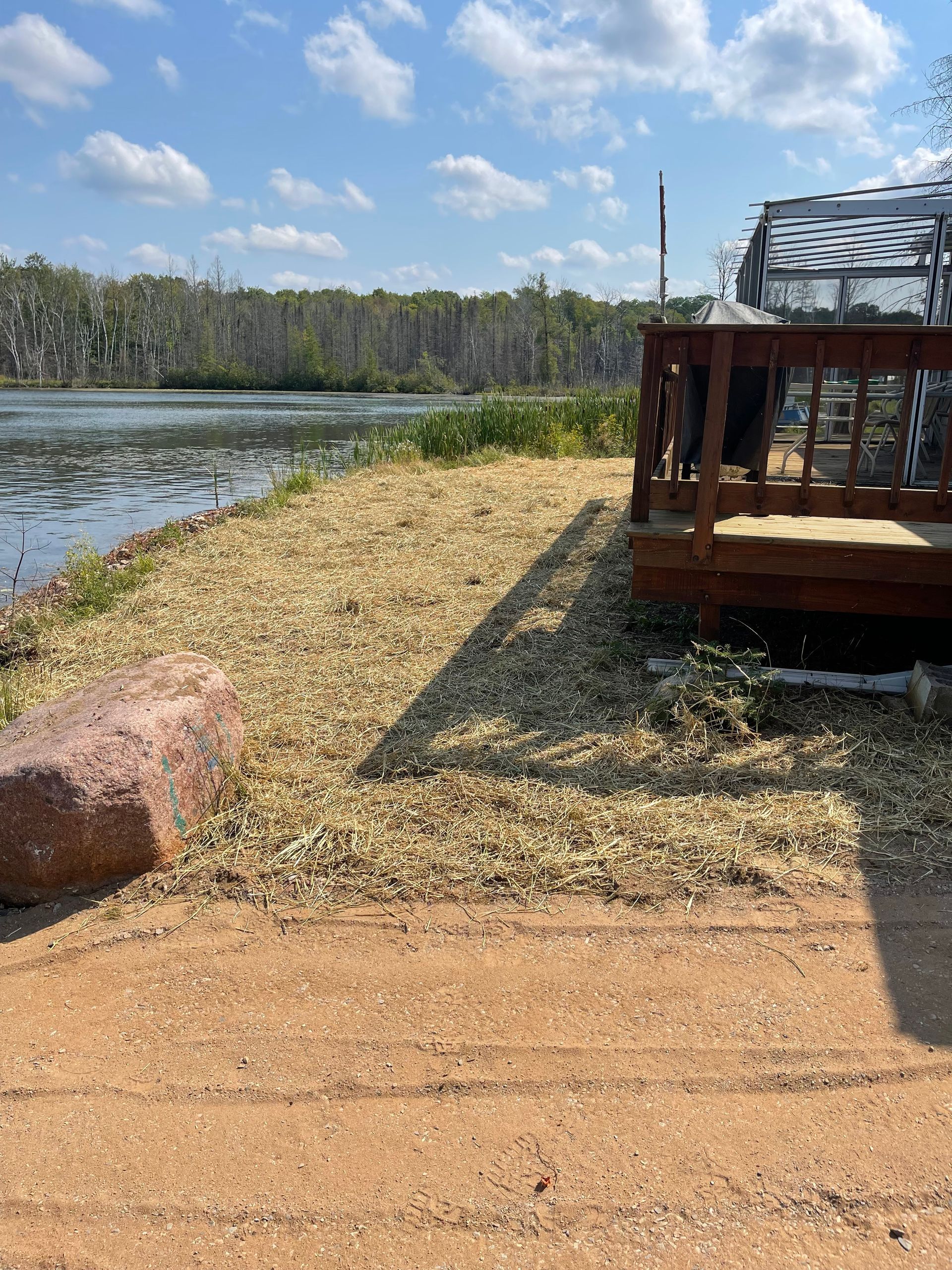 Lakeside view with gravel ground cover, wooden deck, and a large rock in front of water and trees under a sunny sky.