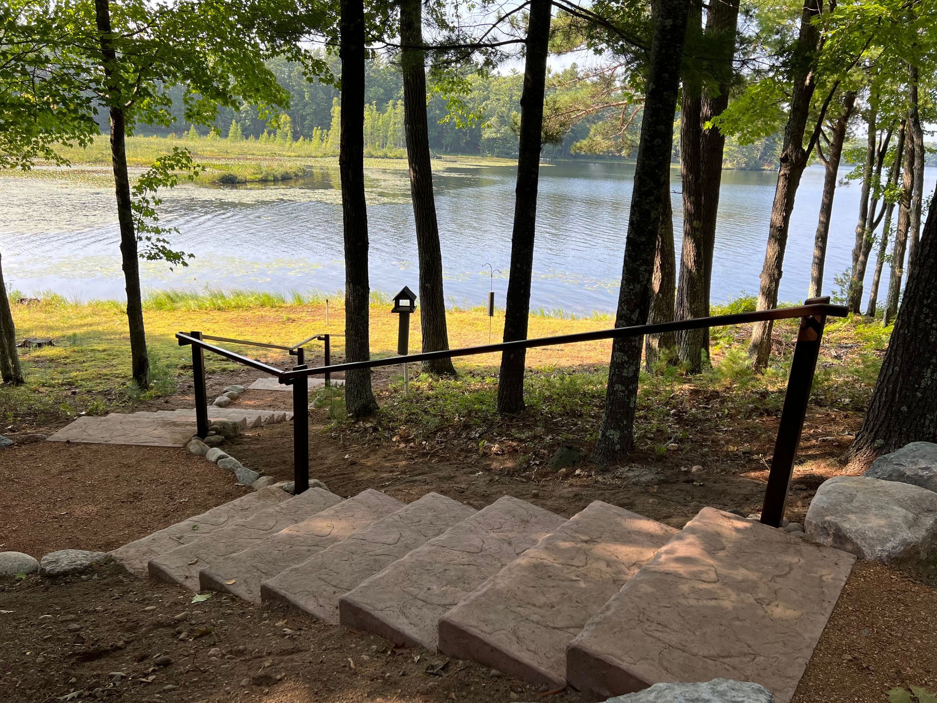 Concrete steps with a black handrail lead to a lake view; trees surround.