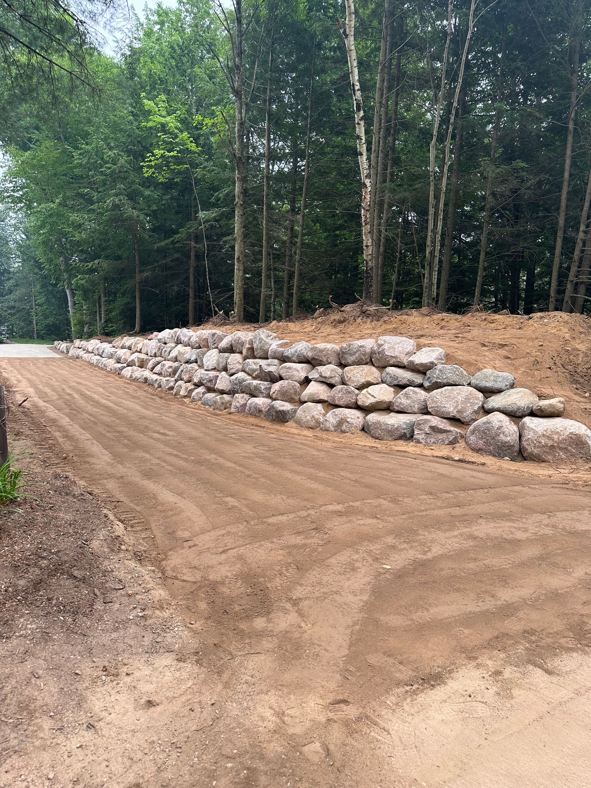 Dirt road alongside a stacked stone retaining wall. Trees in the background.