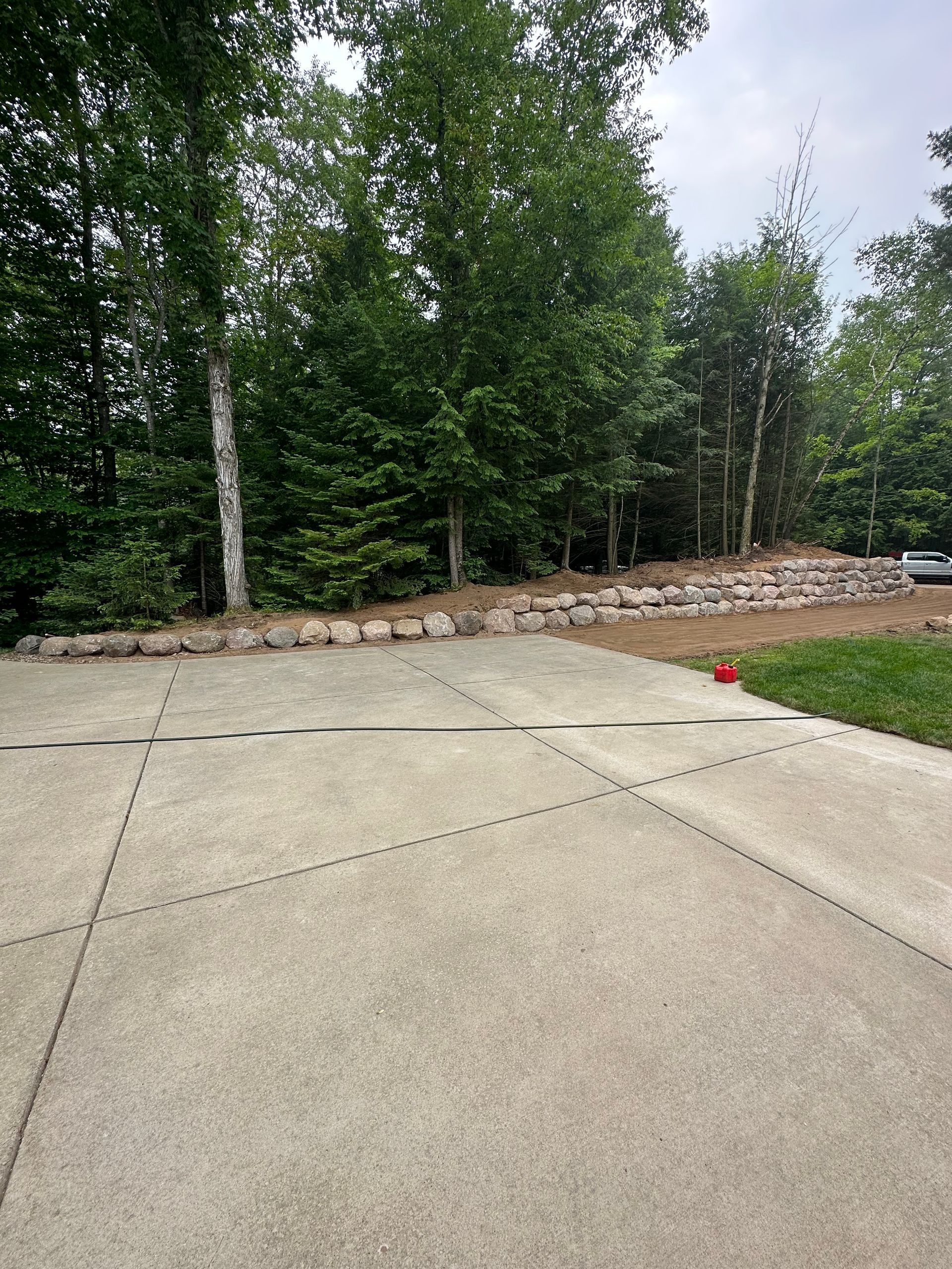 Concrete driveway leading toward trees and a rock wall.