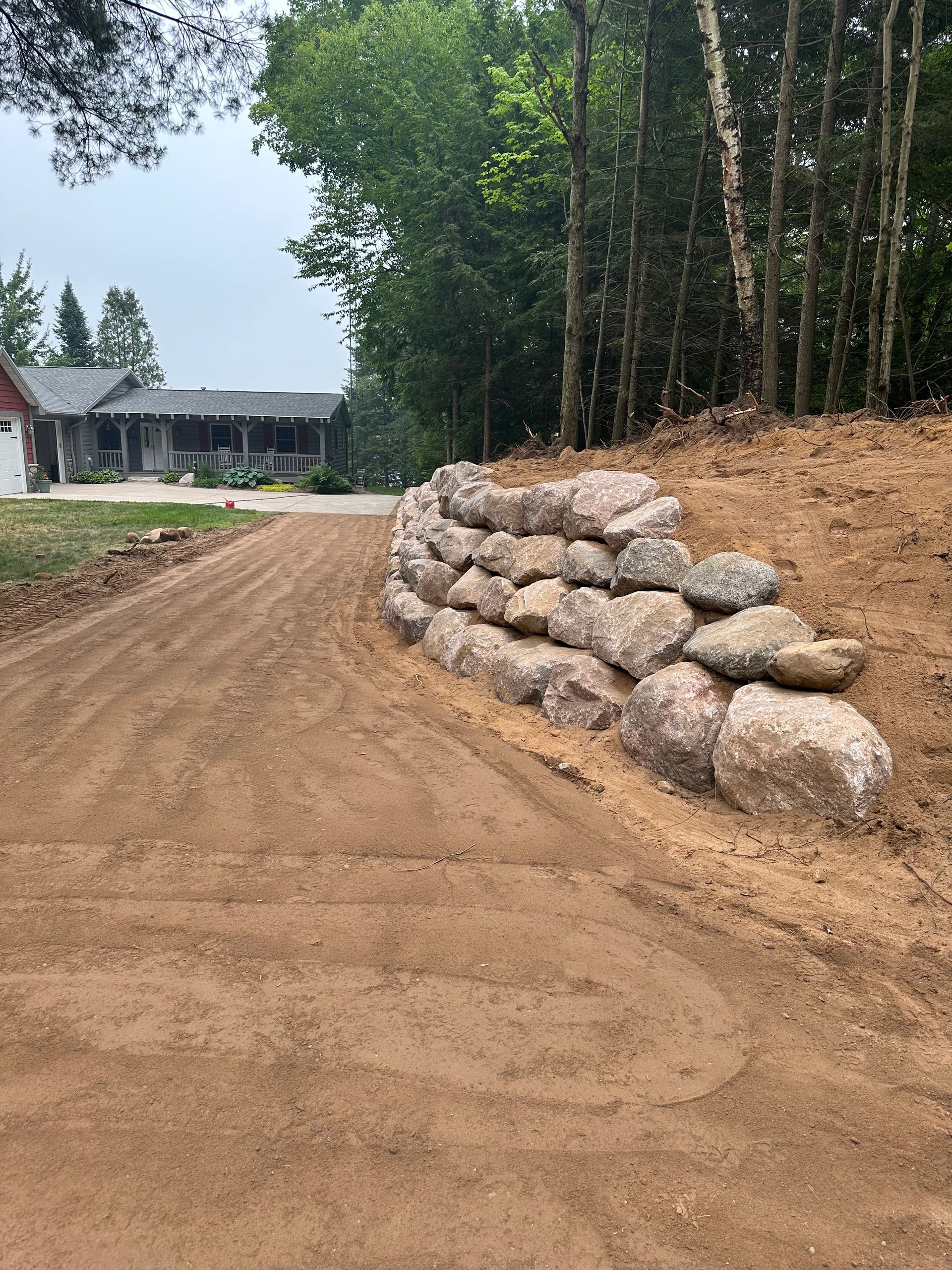 Gravel driveway ascending to a house with a stone retaining wall. Trees in the background, overcast sky.