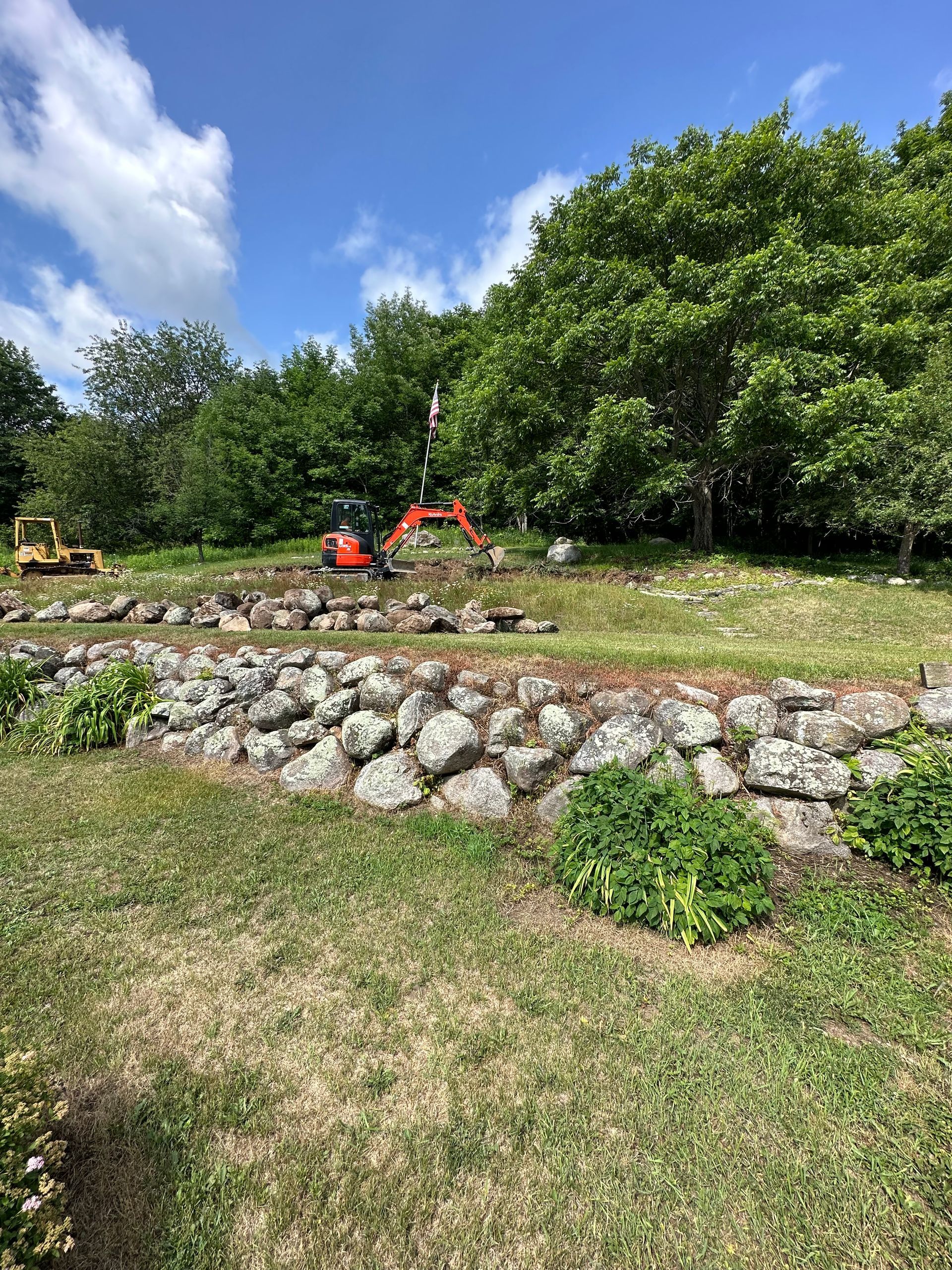Construction site with two excavators. Stone wall in foreground, trees and blue sky.