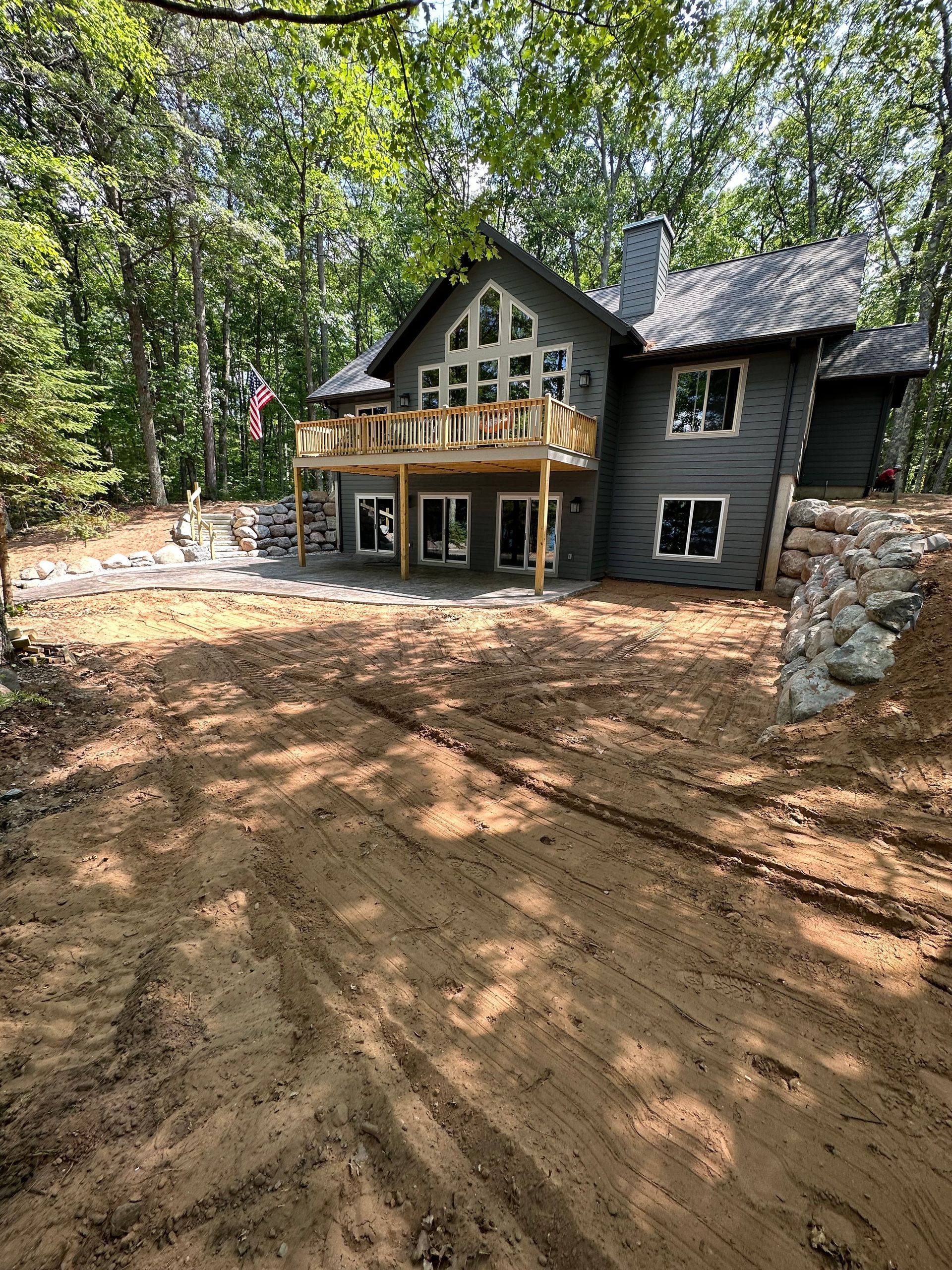 Gray cabin-style home in a wooded area, with a wooden deck and retaining walls, and visible tire tracks.