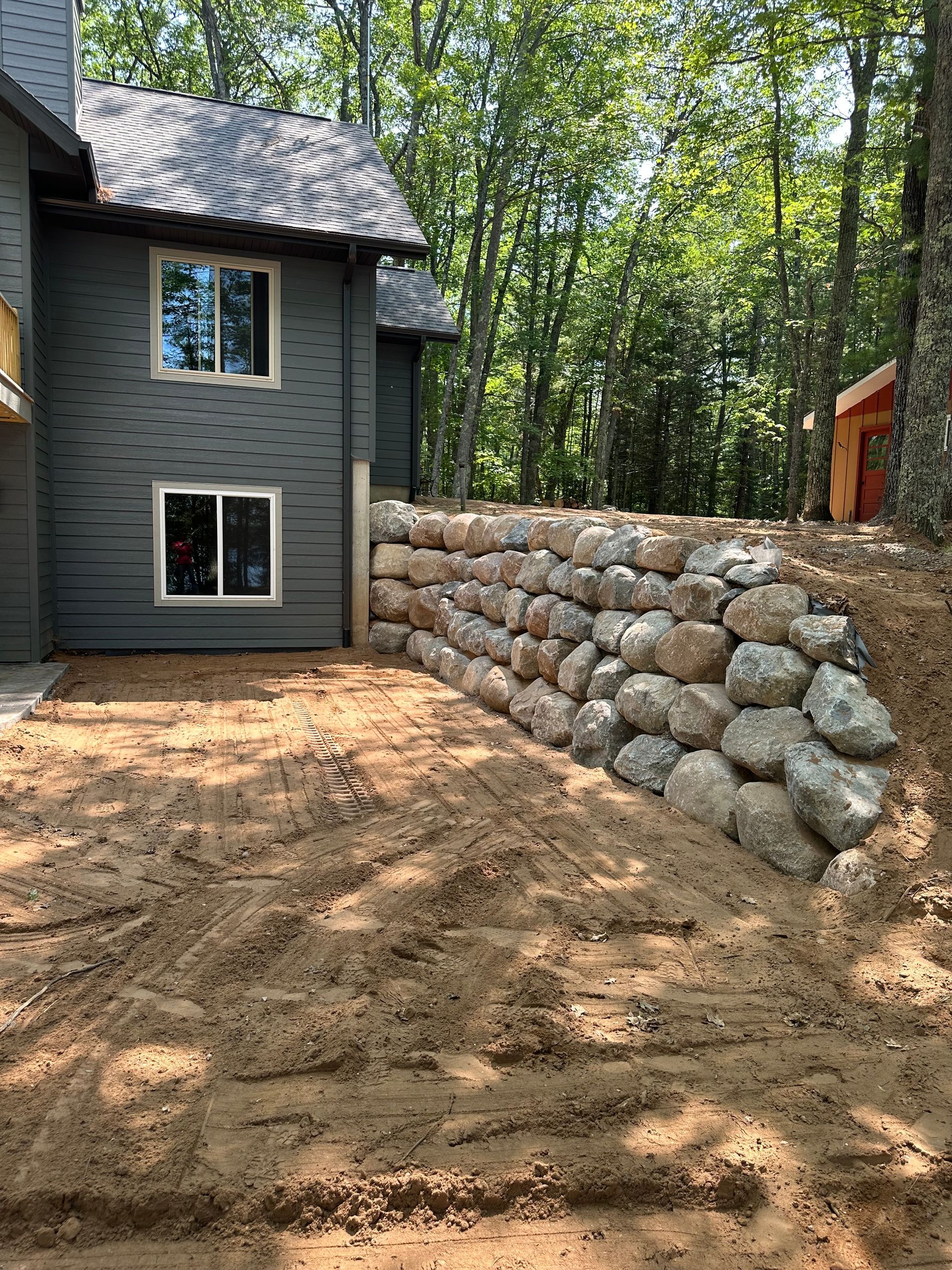 Gray house next to a rock retaining wall, set in dirt, trees in the background.