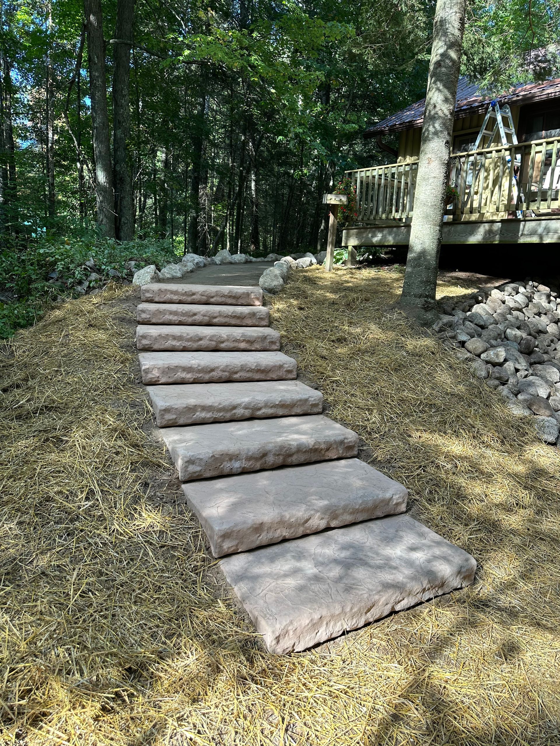 Stone steps leading upwards through a wooded area to a wooden deck. Mulch and rocks border the steps.