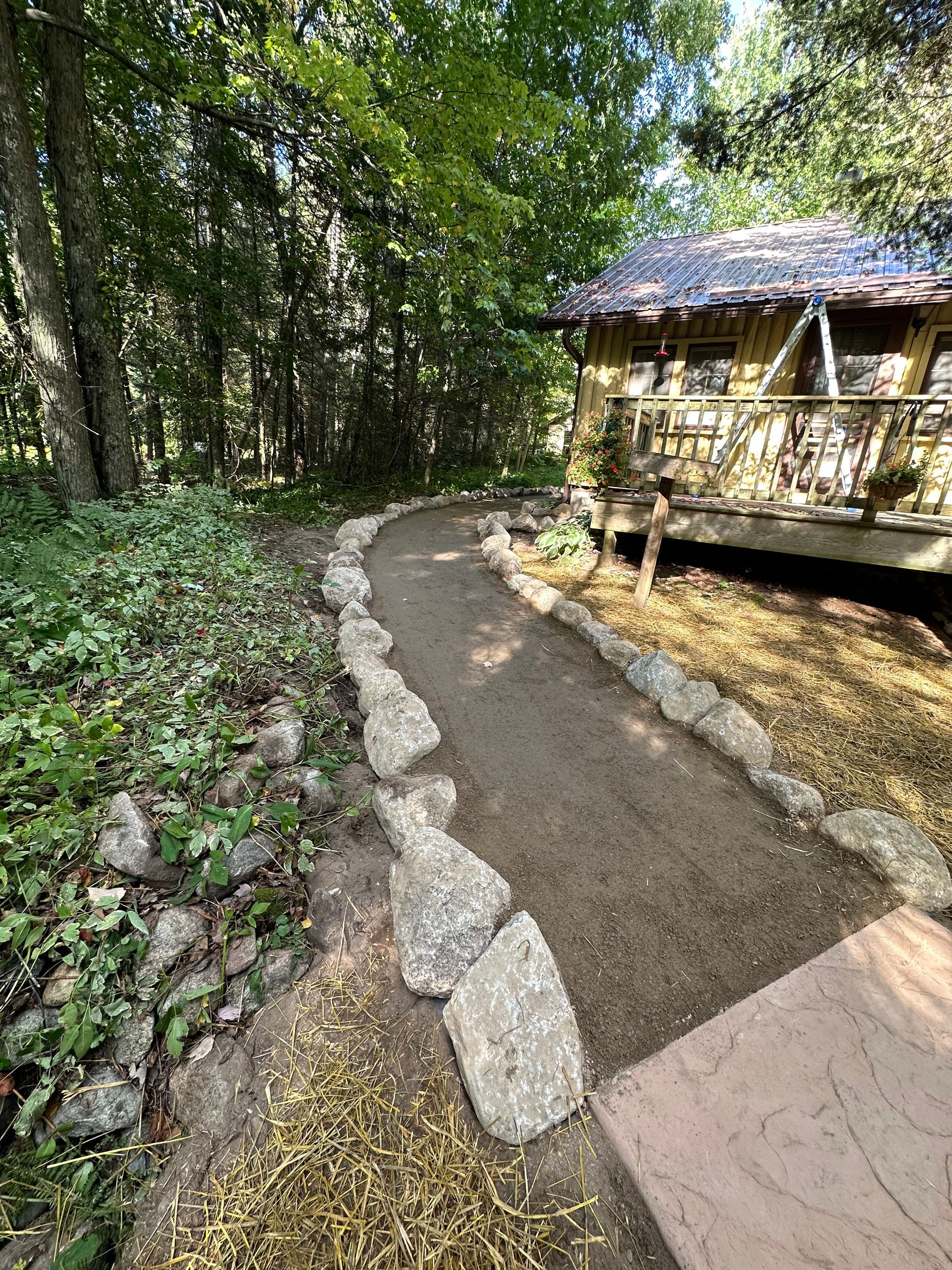 A gravel path winds through a forest, leading to a cabin with a thatched roof.