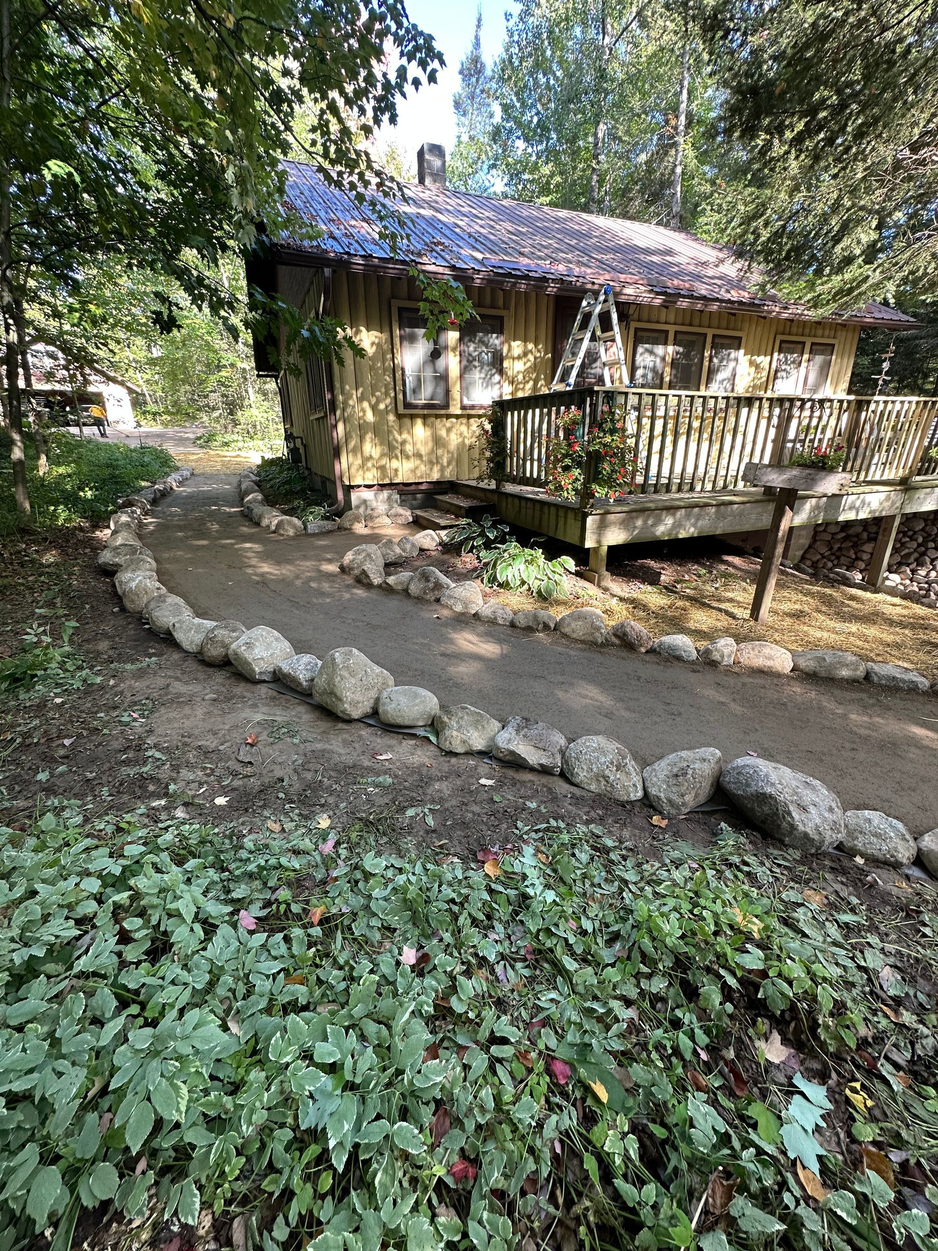 A small, yellow cottage with a wooden deck and a rock-lined pathway in a wooded area.