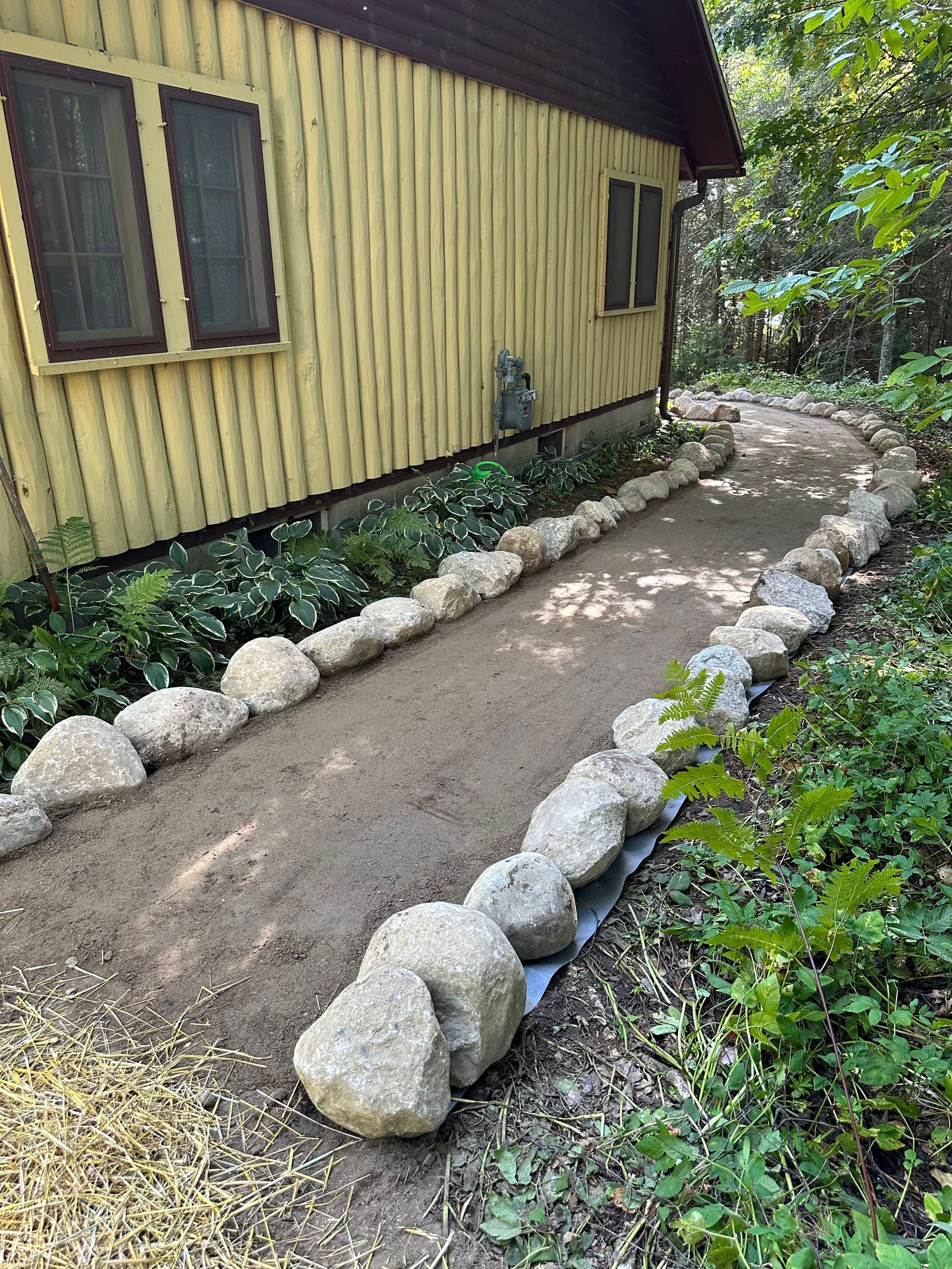 A gravel path lined with rocks curves alongside a yellow building with green foliage.