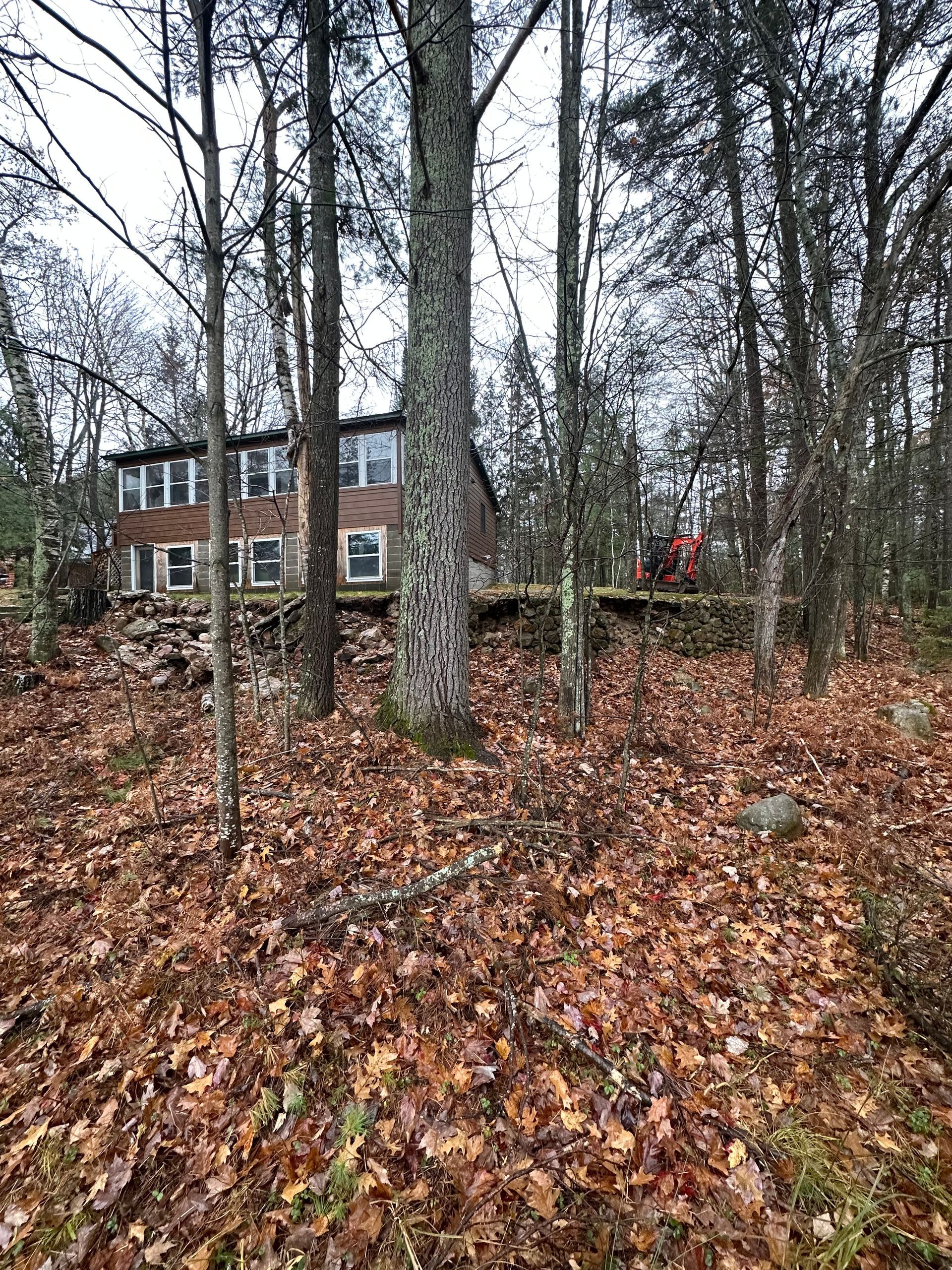 A two-story brick house in the woods, visible through trees and covered in fallen leaves.