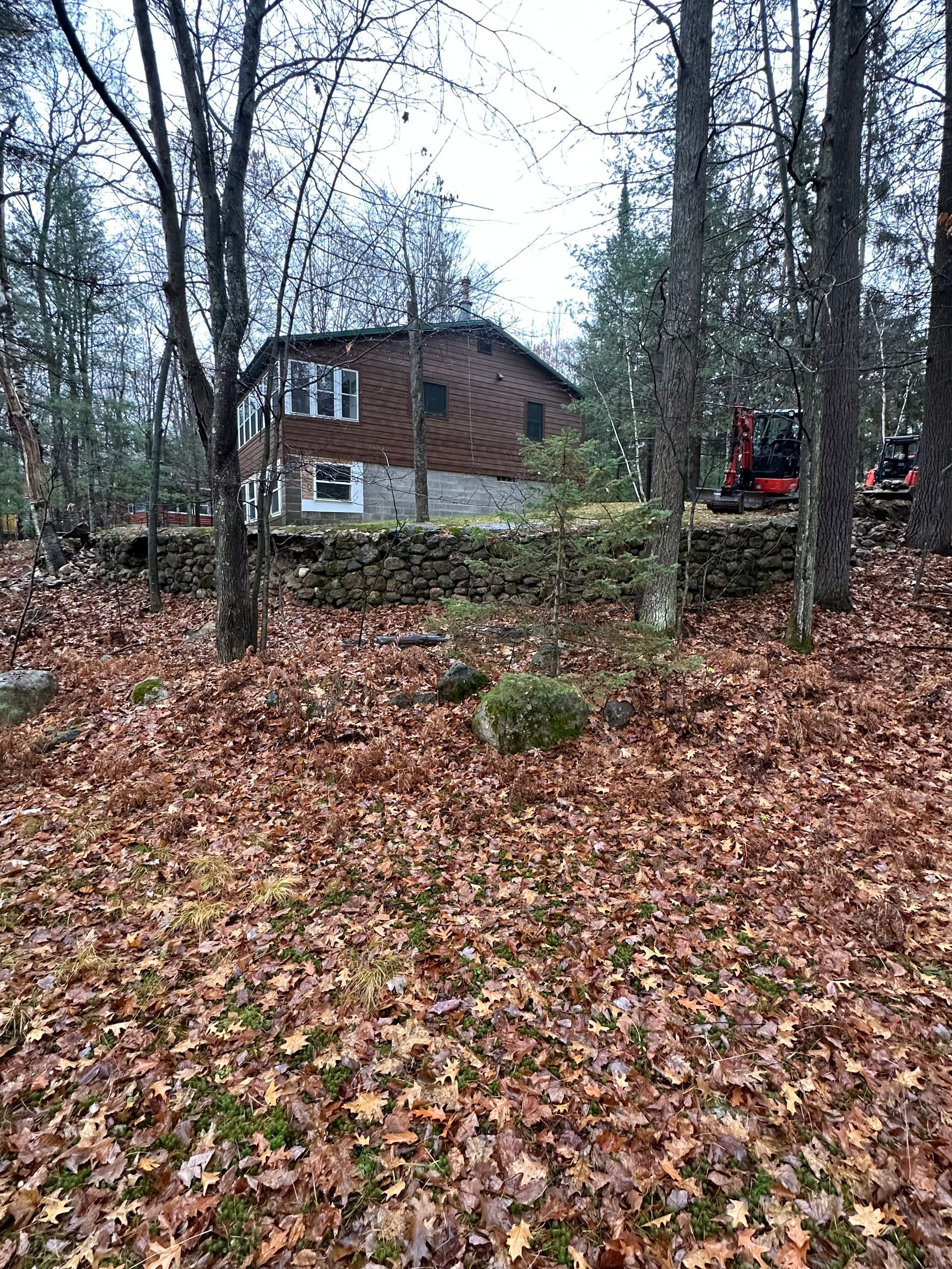 Two-story house with stone foundation set into hillside, surrounded by trees and fallen leaves.