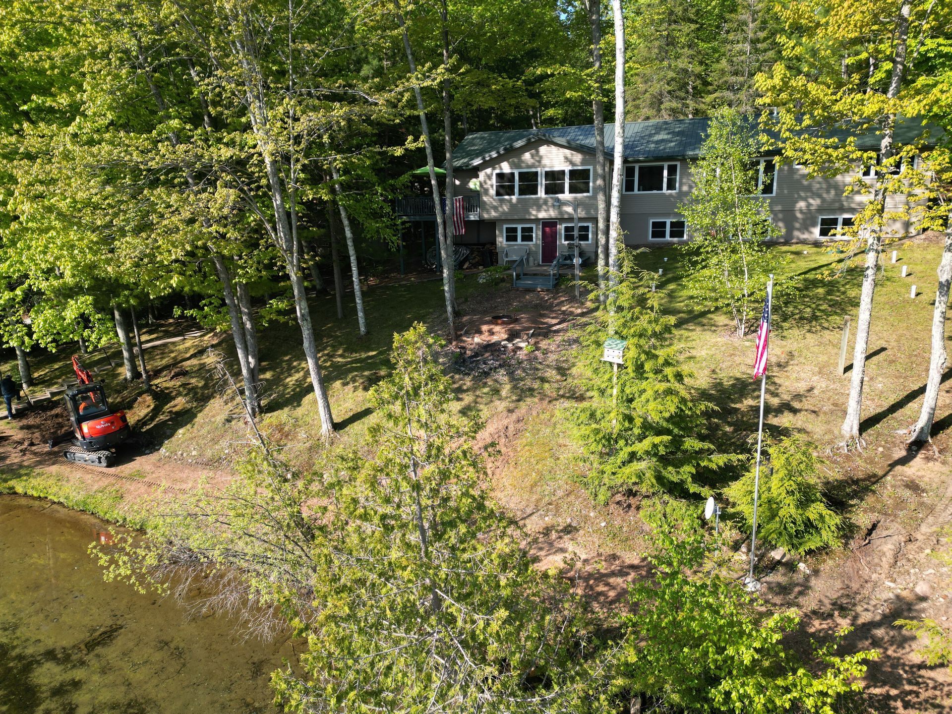 House nestled in trees, next to a pond. Red door, small excavator, American flag.