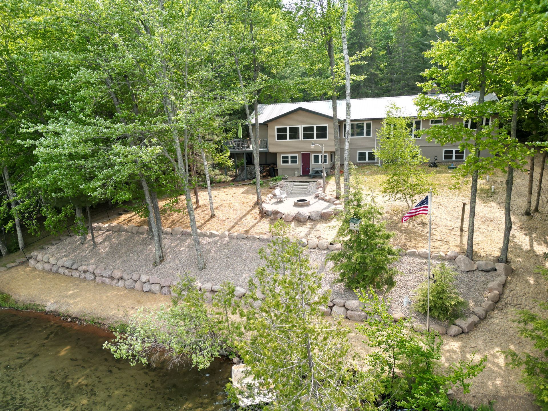 Cottage nestled in trees near a lake. Fire pit and American flag in yard.