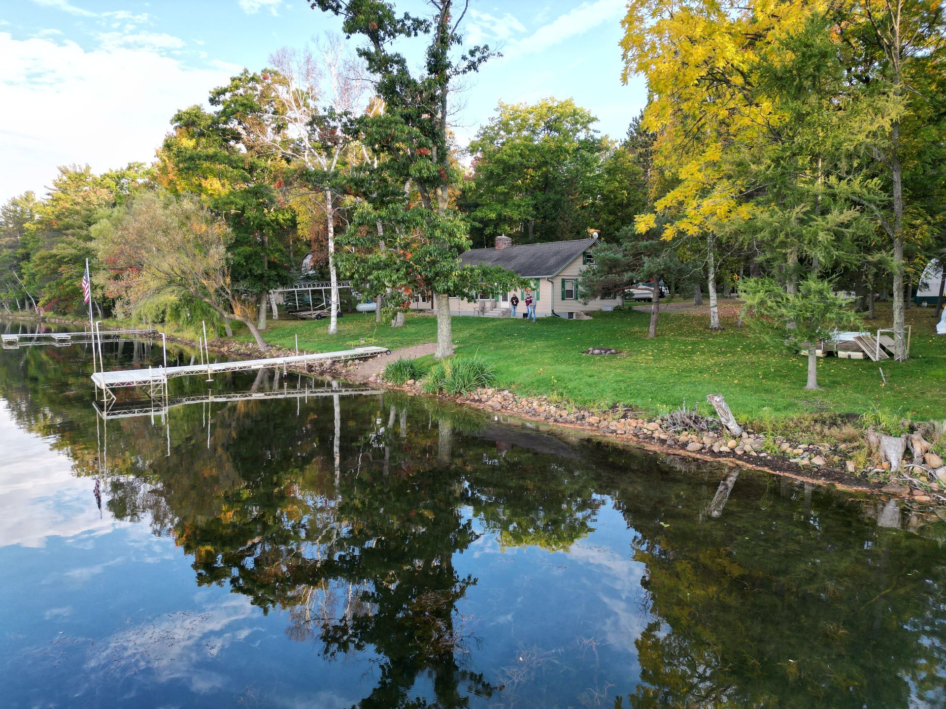 Waterfront cottage with a dock, trees with colorful leaves, and reflections in the calm water.