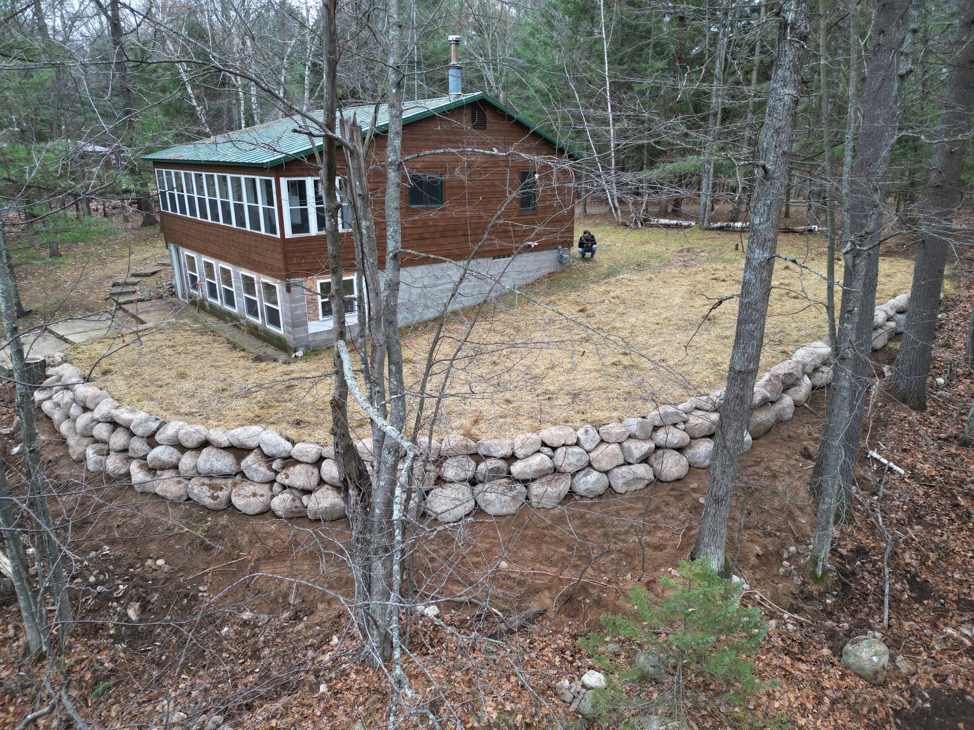 Cabin with stone wall in a wooded area. Brown building with many windows, green roof.