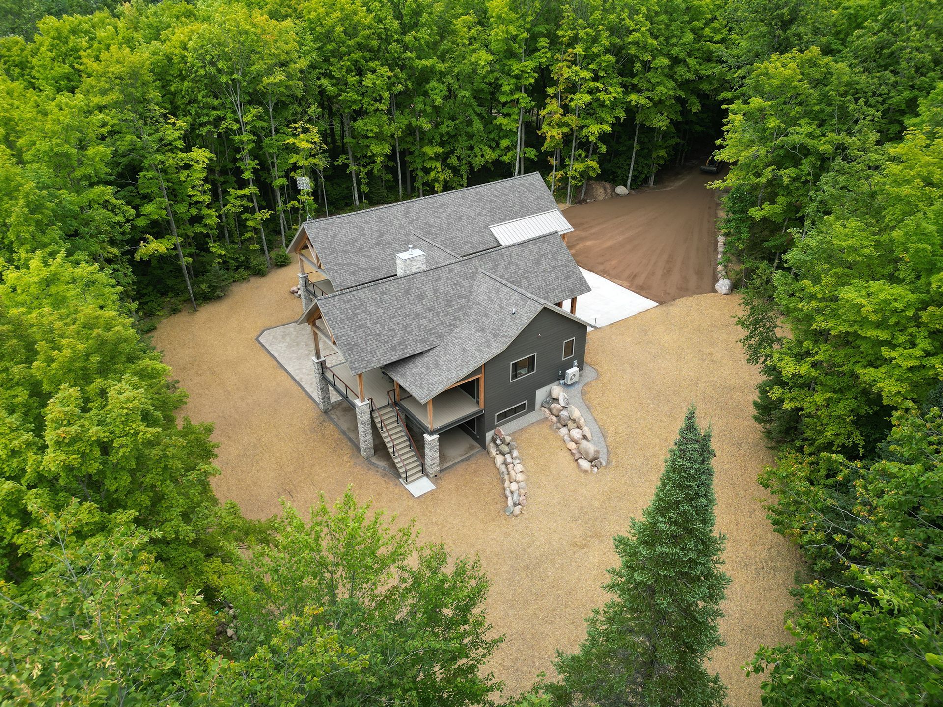 Aerial view of a gray-roofed cabin surrounded by trees and gravel, with a small cleared driveway.