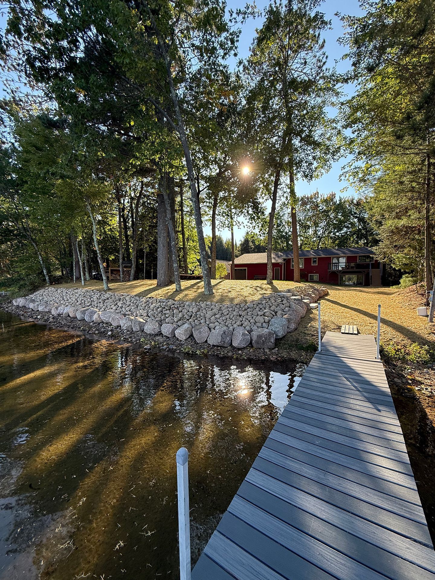 Lake scene with dock, trees, and small red building. Sun shining.