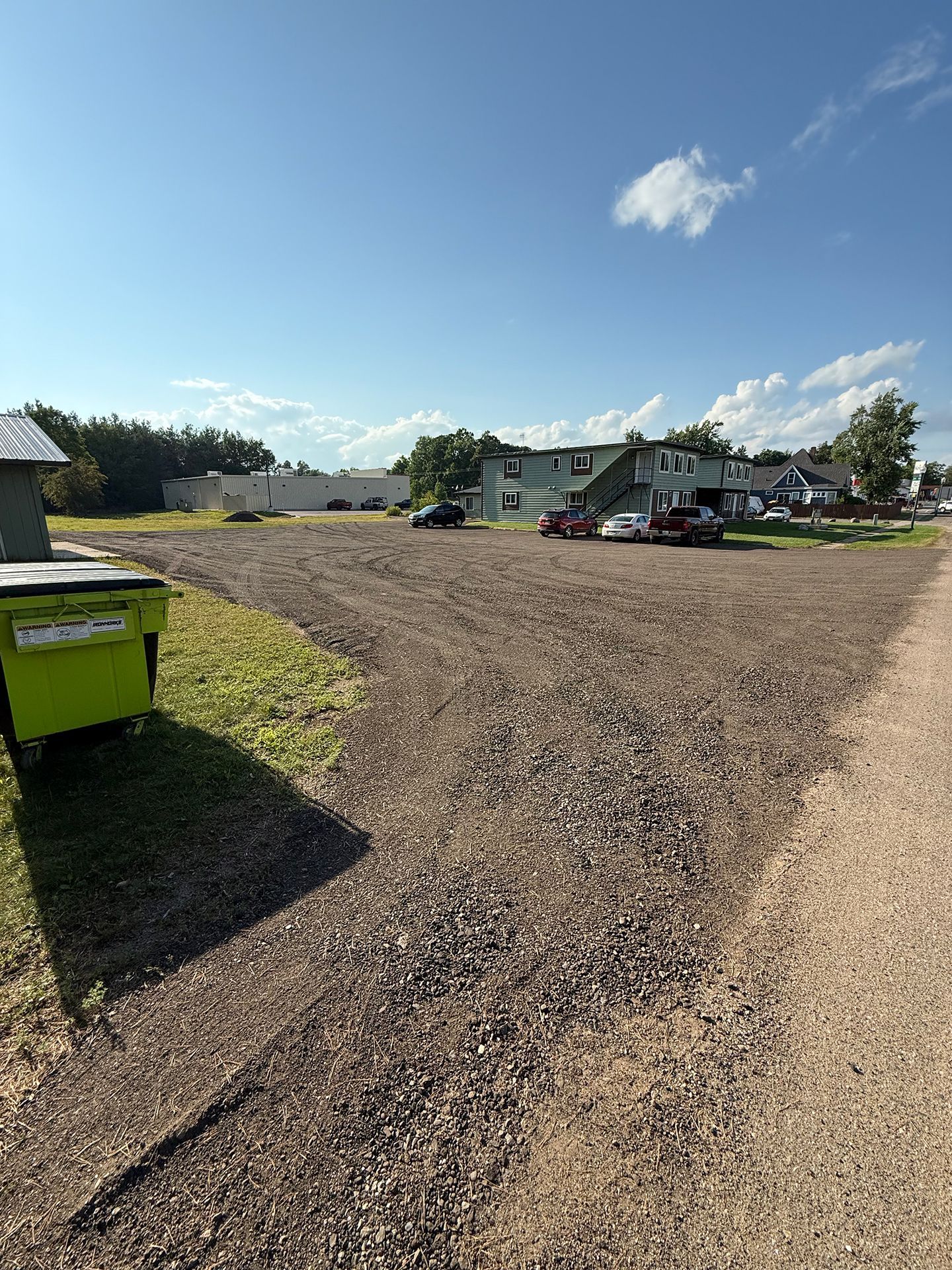 Gravel parking lot with green dumpster on the left, building and cars in the background under a blue sky.