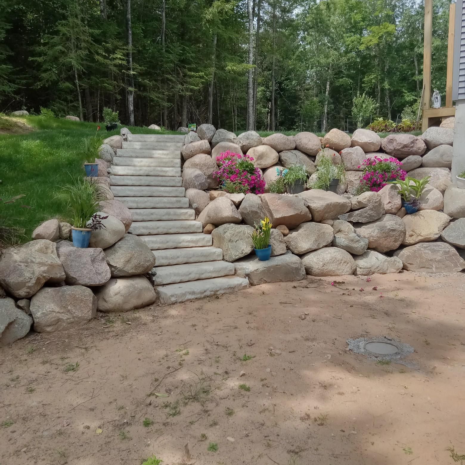 Stone steps leading up a rock retaining wall with potted flowers and trees in the background.
