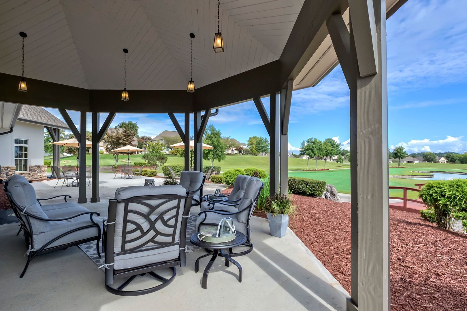 Outdoor patio with seating, overlooking a golf course. Gray furniture, red mulch, and blue sky.