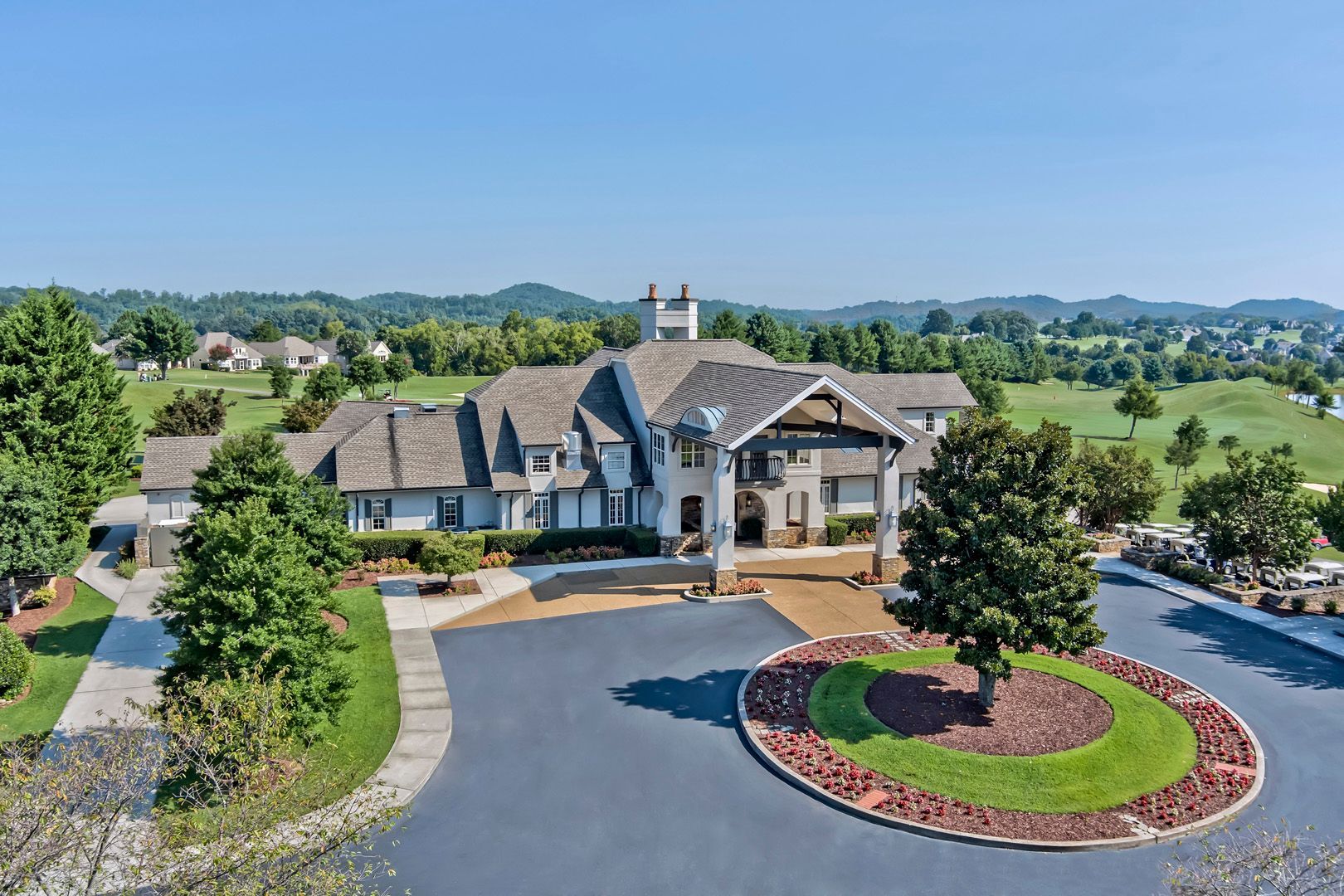 Large gray and white house with a circular driveway and manicured landscaping, set against a green, hilly backdrop.
