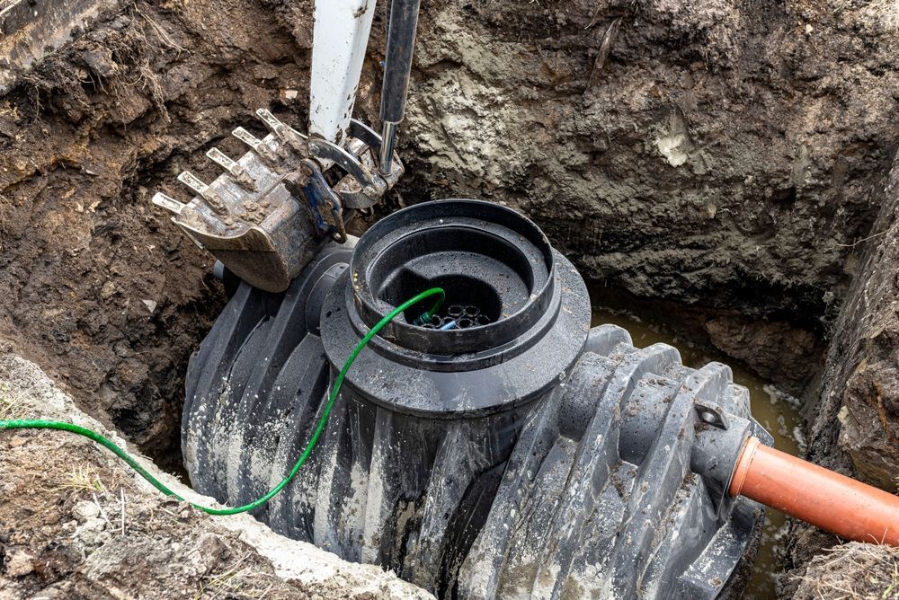 A backhoe excavating a sewage tank in a dirt pit. The tank is black, with an orange pipe attached.