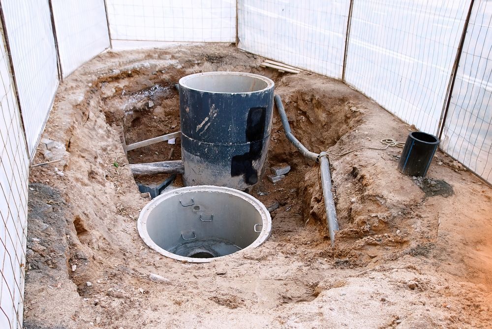 Excavated area with two open, cylindrical containers, one taller with cutouts, surrounded by dirt and plumbing.
