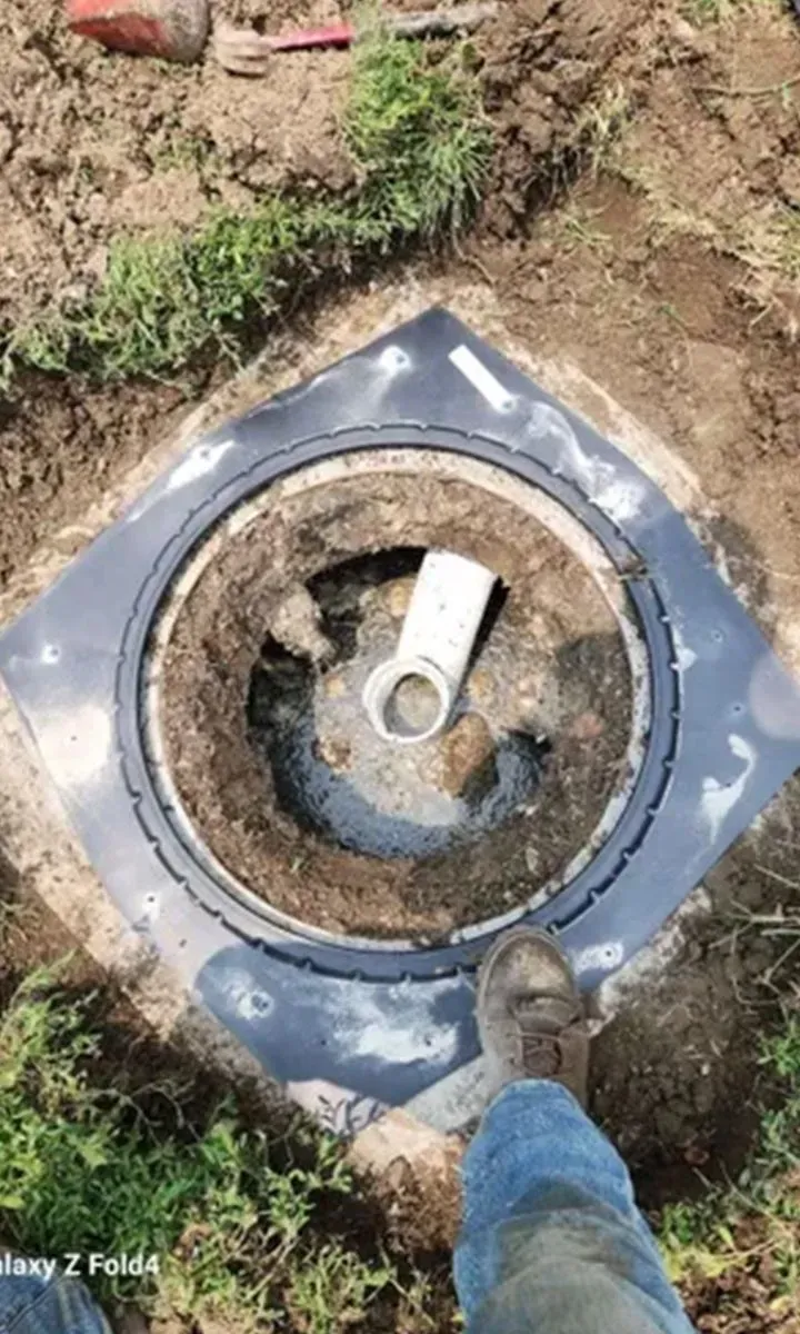 Open well with white pipe, surrounded by dirt and black plastic square; a person's foot visible.