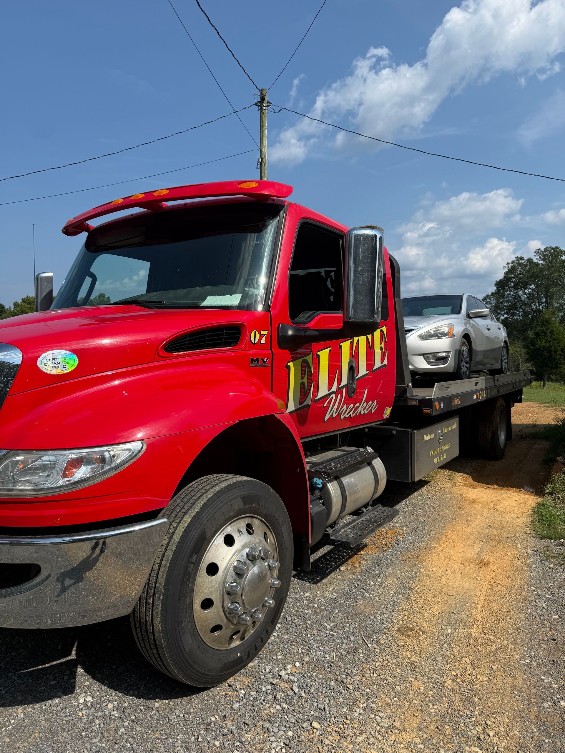 Red tow truck hauling a silver car on a gravel roadside under a blue sky