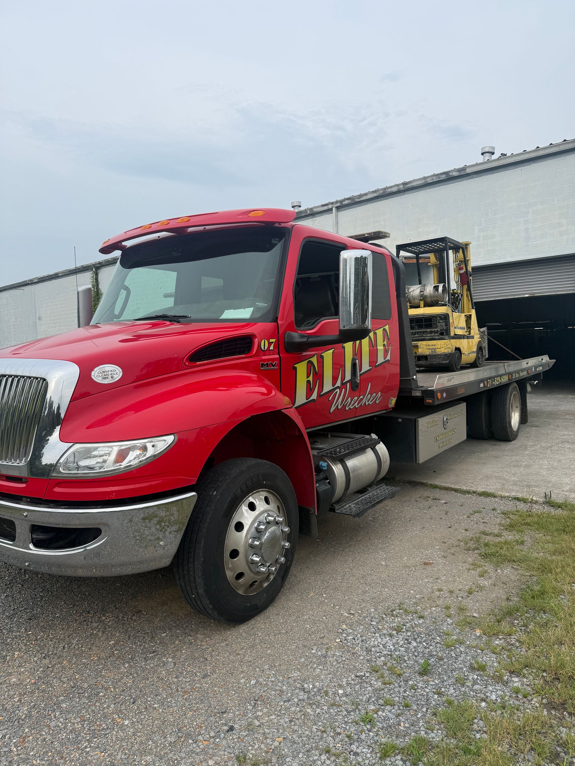 Red tow truck carrying a yellow forklift in a gravel lot beside a warehouse