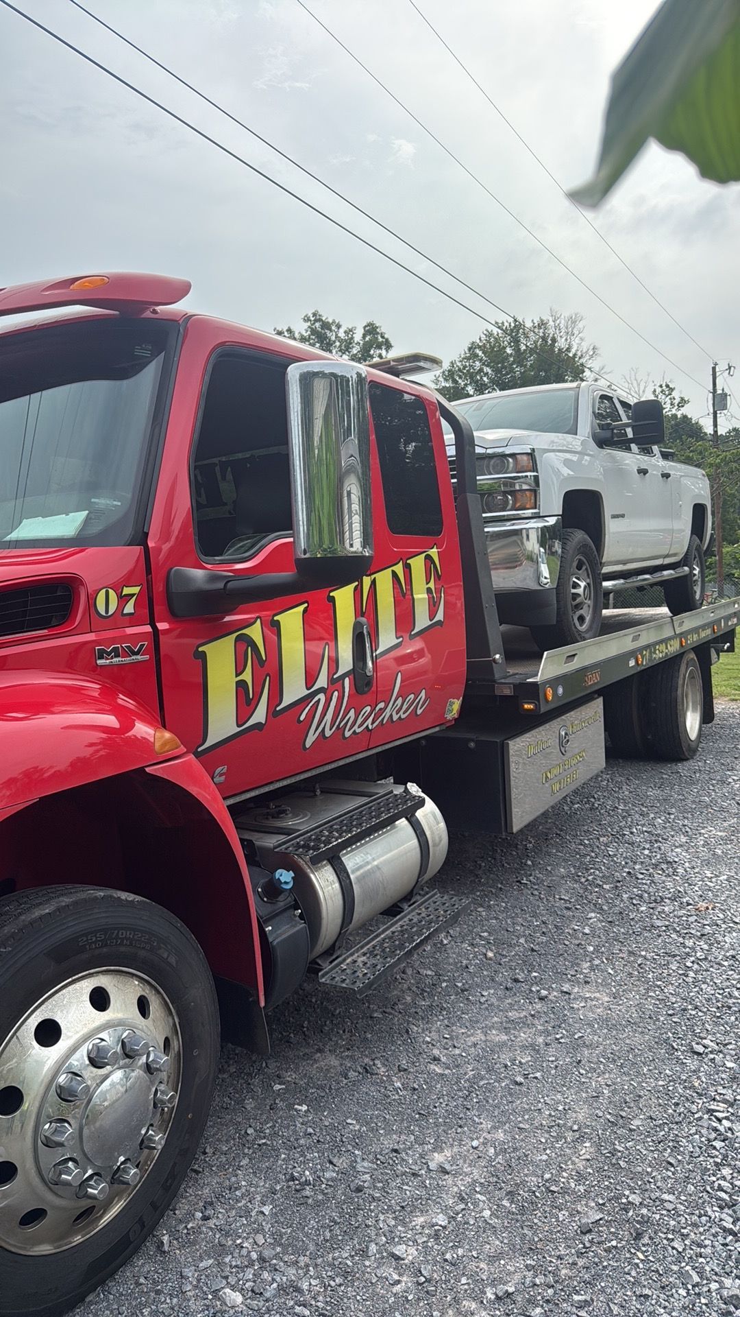 Red tow truck with ELITE logo hauling a white SUV on a gravel roadside under cloudy skies